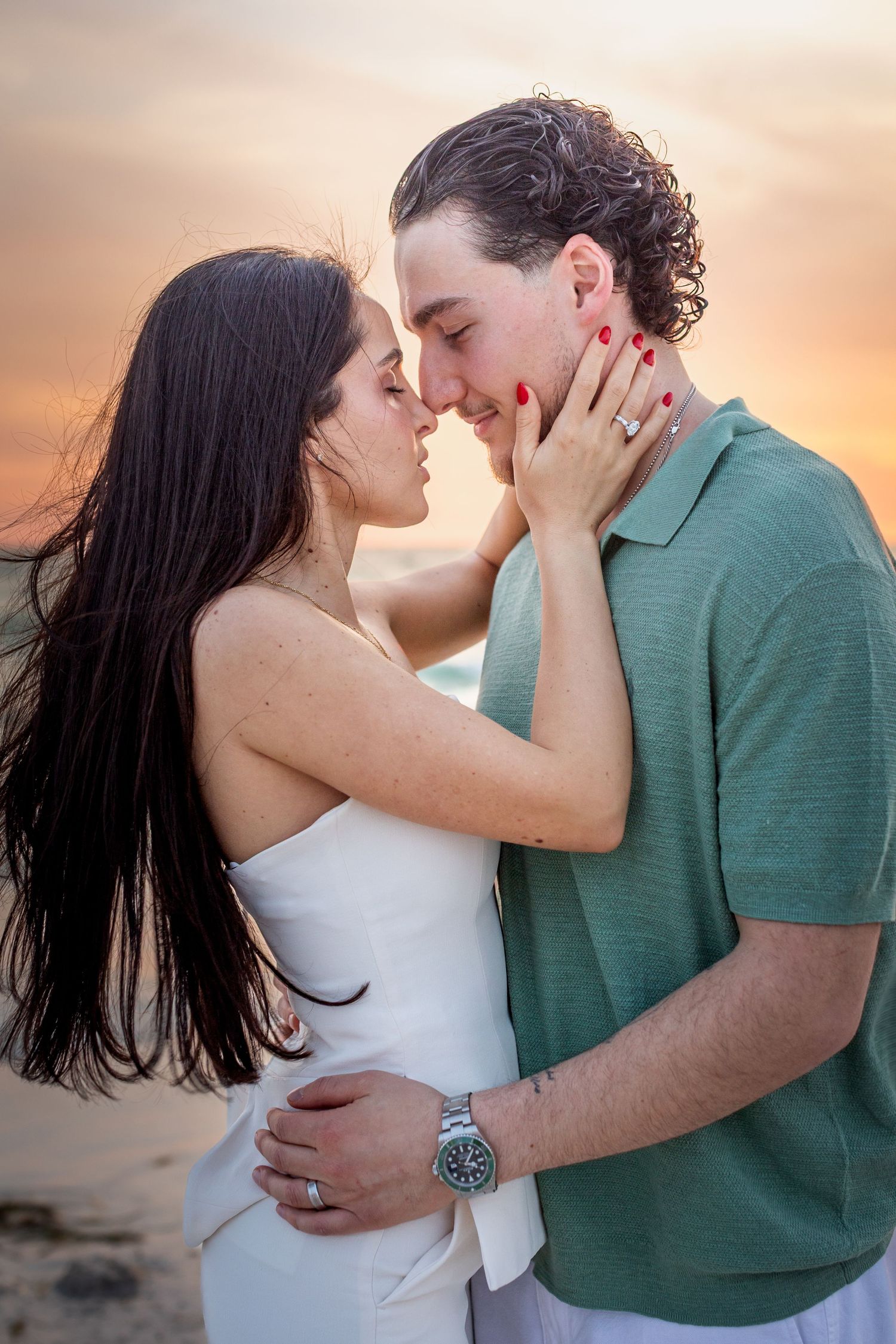 Couple sharing a romantic almost-kiss at sunset on the beach, woman in white dress, man in teal shirt wearing a watch.