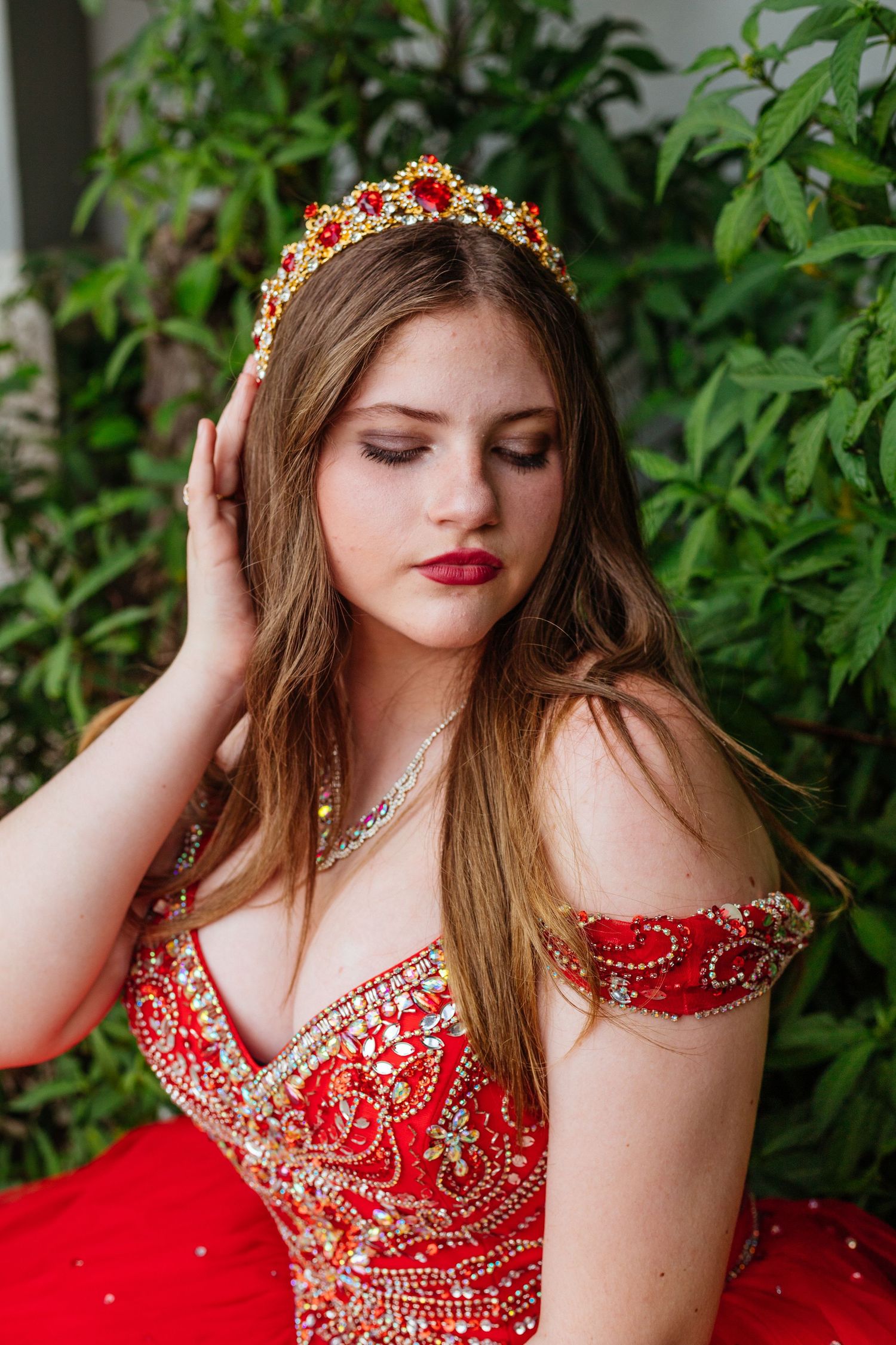 A stunning red ball gown with intricate crystal beading and a gold crown photographed against lush greenery.