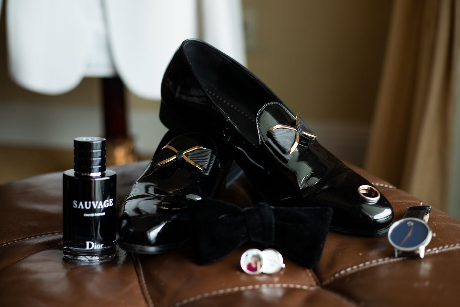 Black patent leather loafers with Dior Sauvage cologne, cufflinks, bow tie, and a watch arranged on brown leather surface.