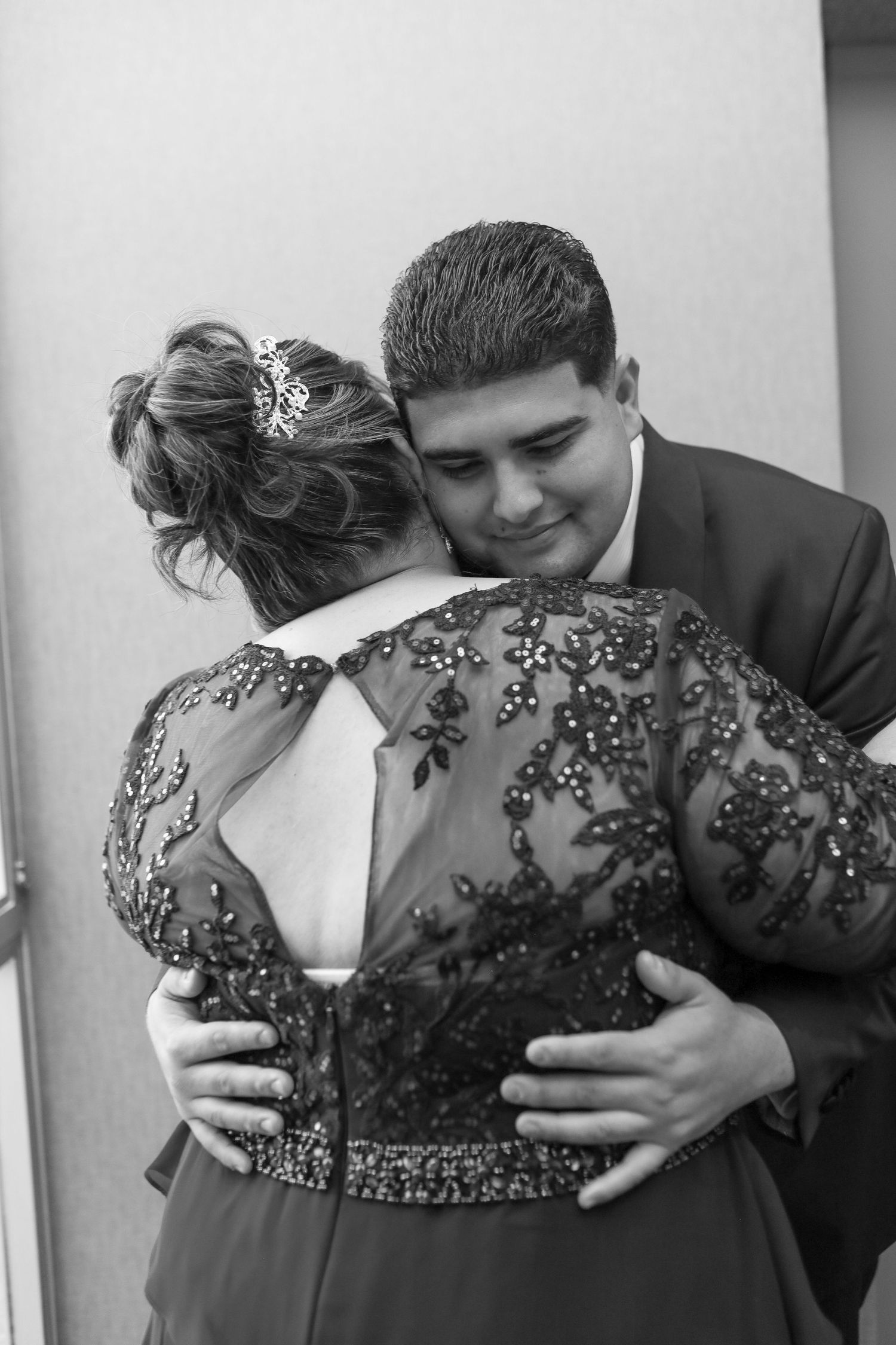 Black and white photo of a young couple embracing, woman in elegant beaded gown with updo, man in suit smiling warmly.