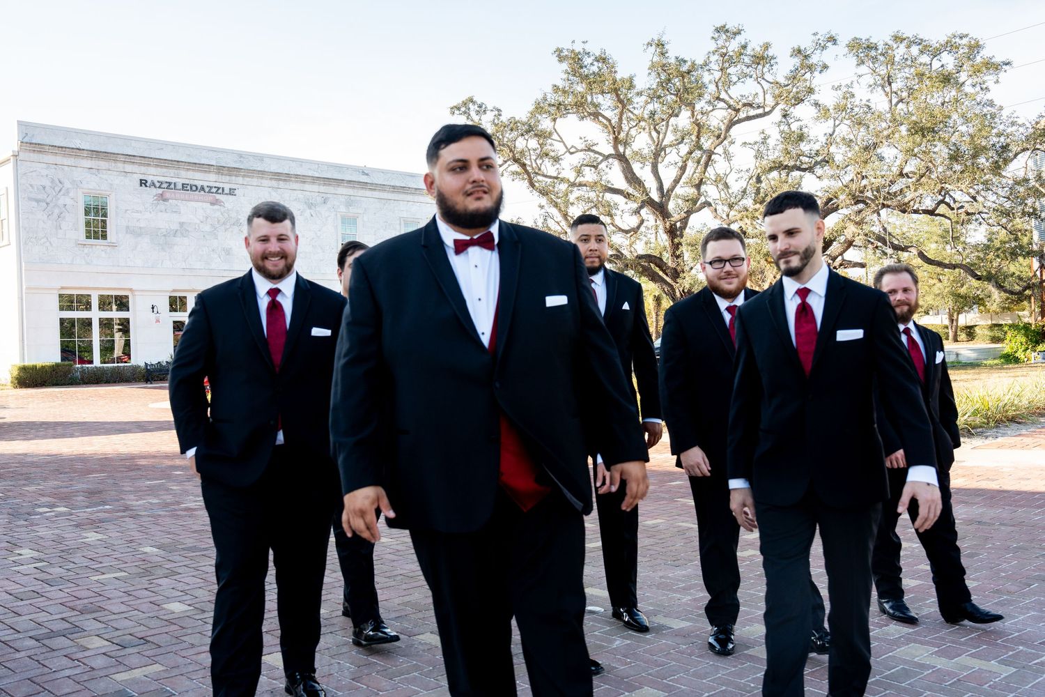 Groom in black tuxedo with red bow tie leads groomsmen in matching suits on brick plaza outside elegant venue.