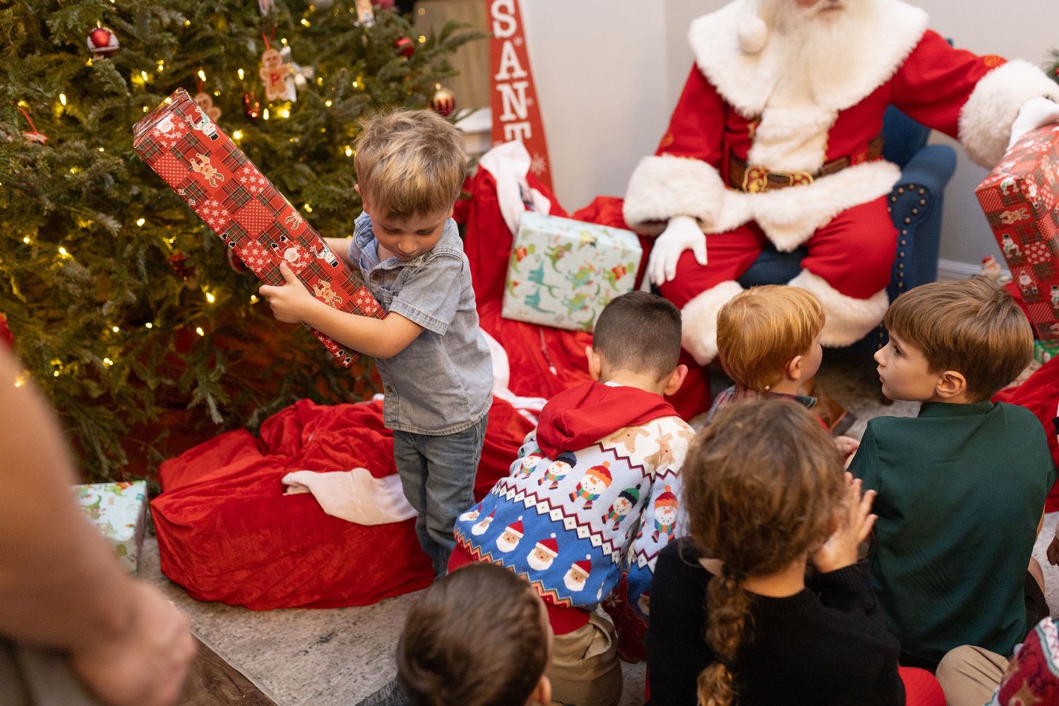 Group of children excitedly interact with Santa Claus near a decorated Christmas tree.