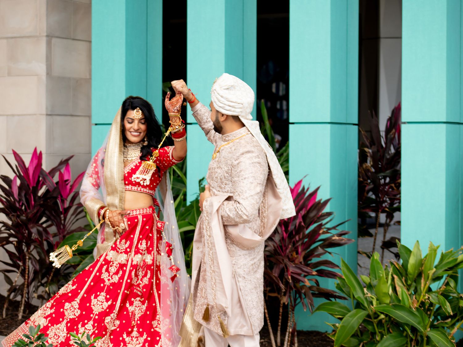 A couple in traditional Indian wedding attire poses against turquoise walls with tropical plants.