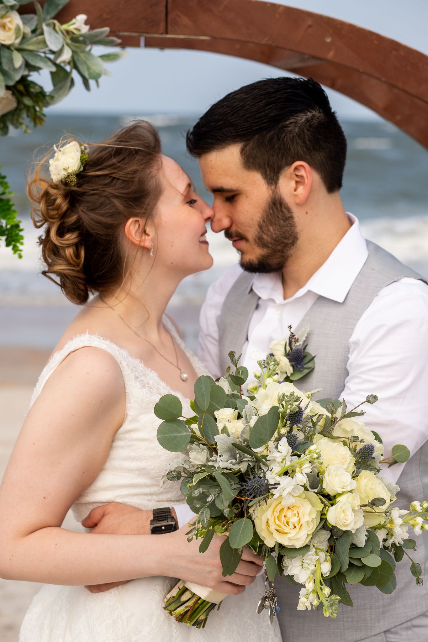 Romantic wedding couple shares intimate moments under a wooden arch with white and yellow floral bouquet.
