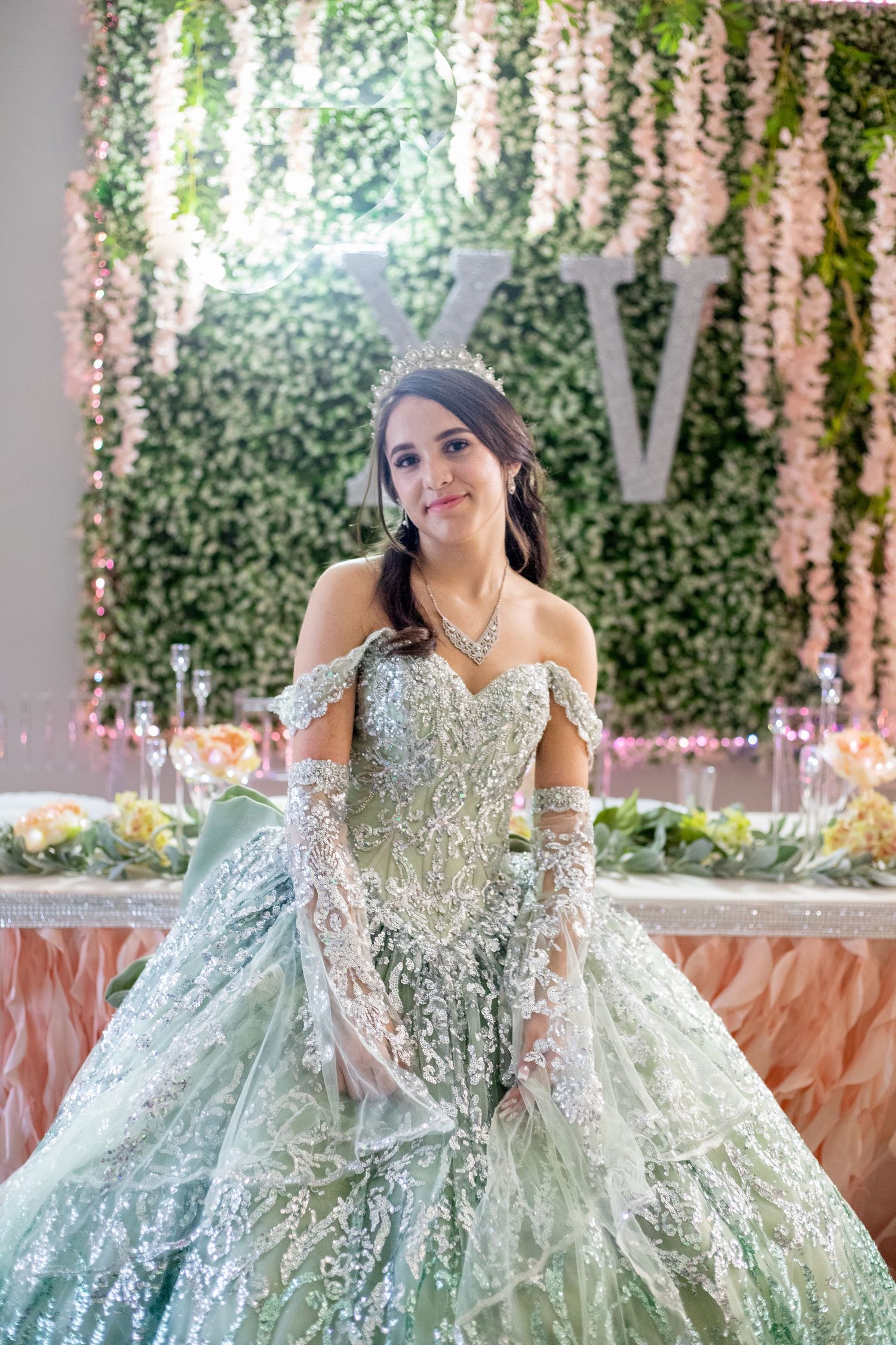 A sparkling mint green ballgown with intricate beading and off-shoulder design against a floral backdrop.