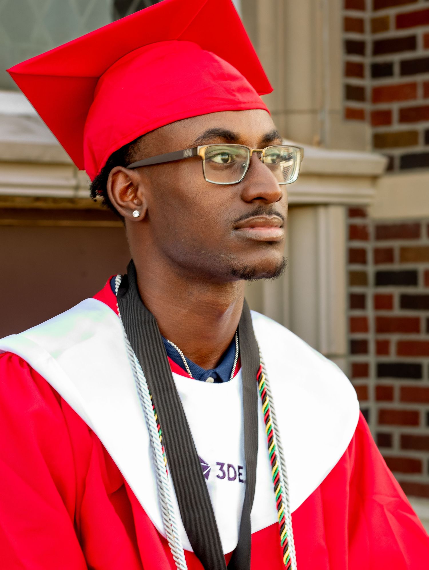 Graduate in red cap and gown with glasses poses in front of brick building.