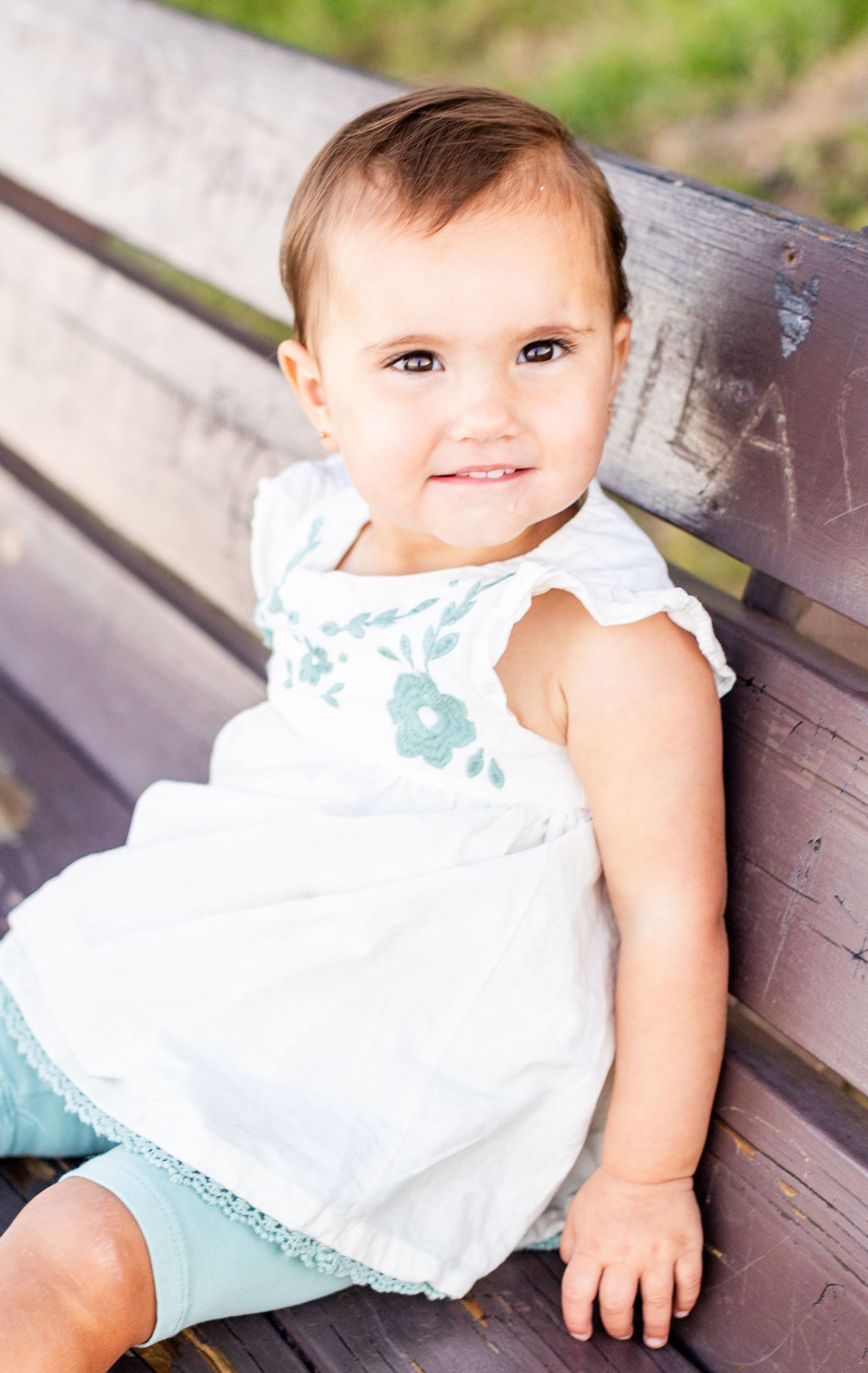 Adorable baby girl in white floral embroidered dress and mint shorts sitting on a wooden bench smiling.