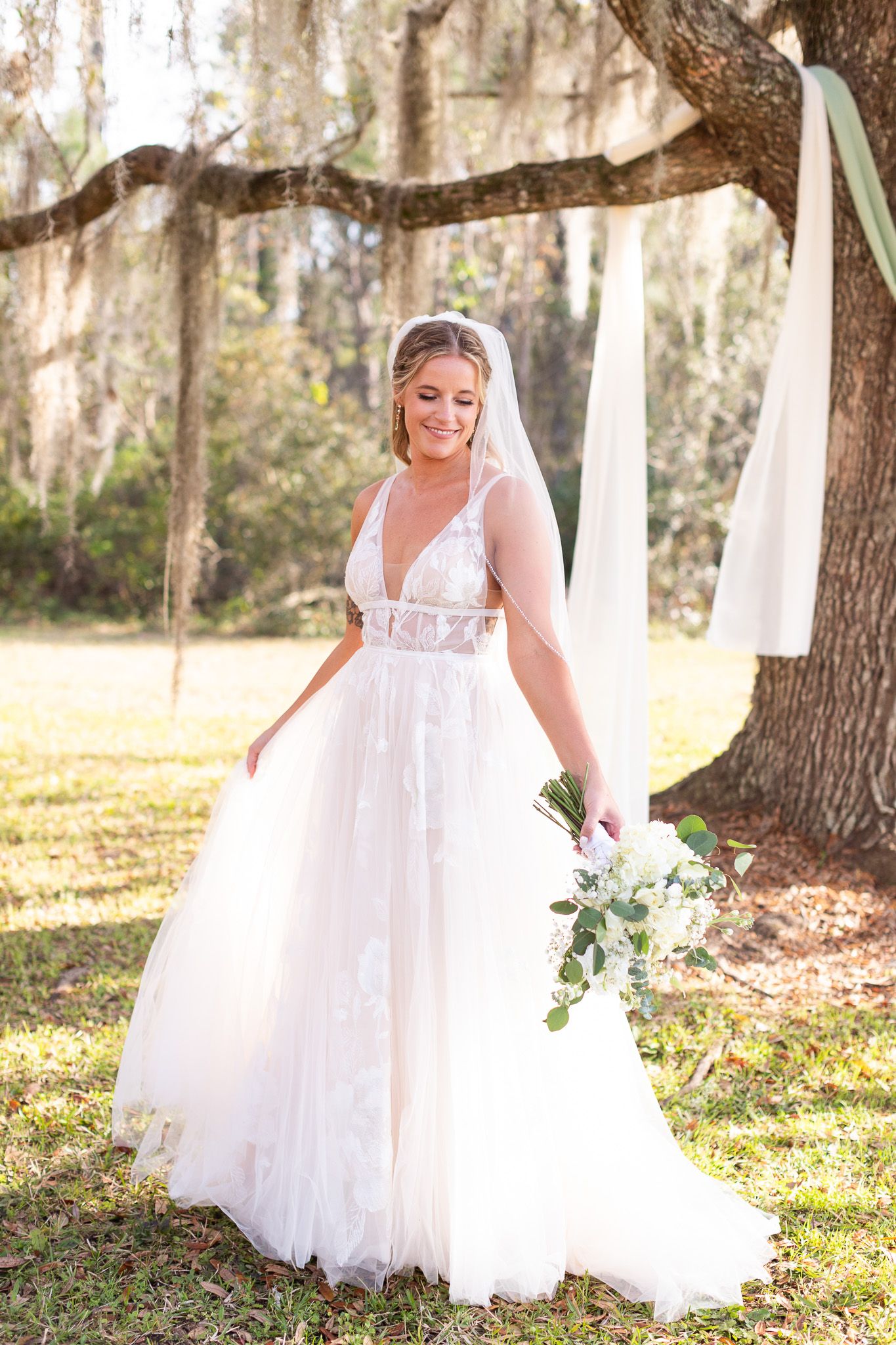 Bride in flowing white dress holds bouquet under mossy oak trees.