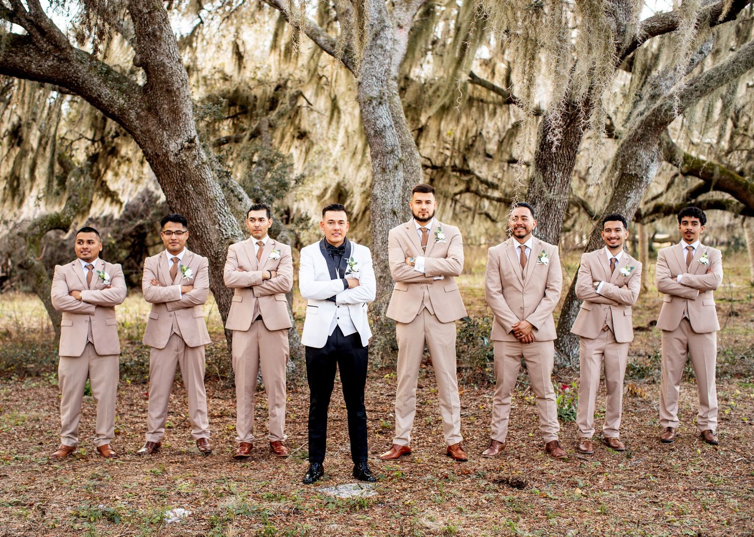 Groomsmen in light beige suits stand in a row under moss-covered trees.