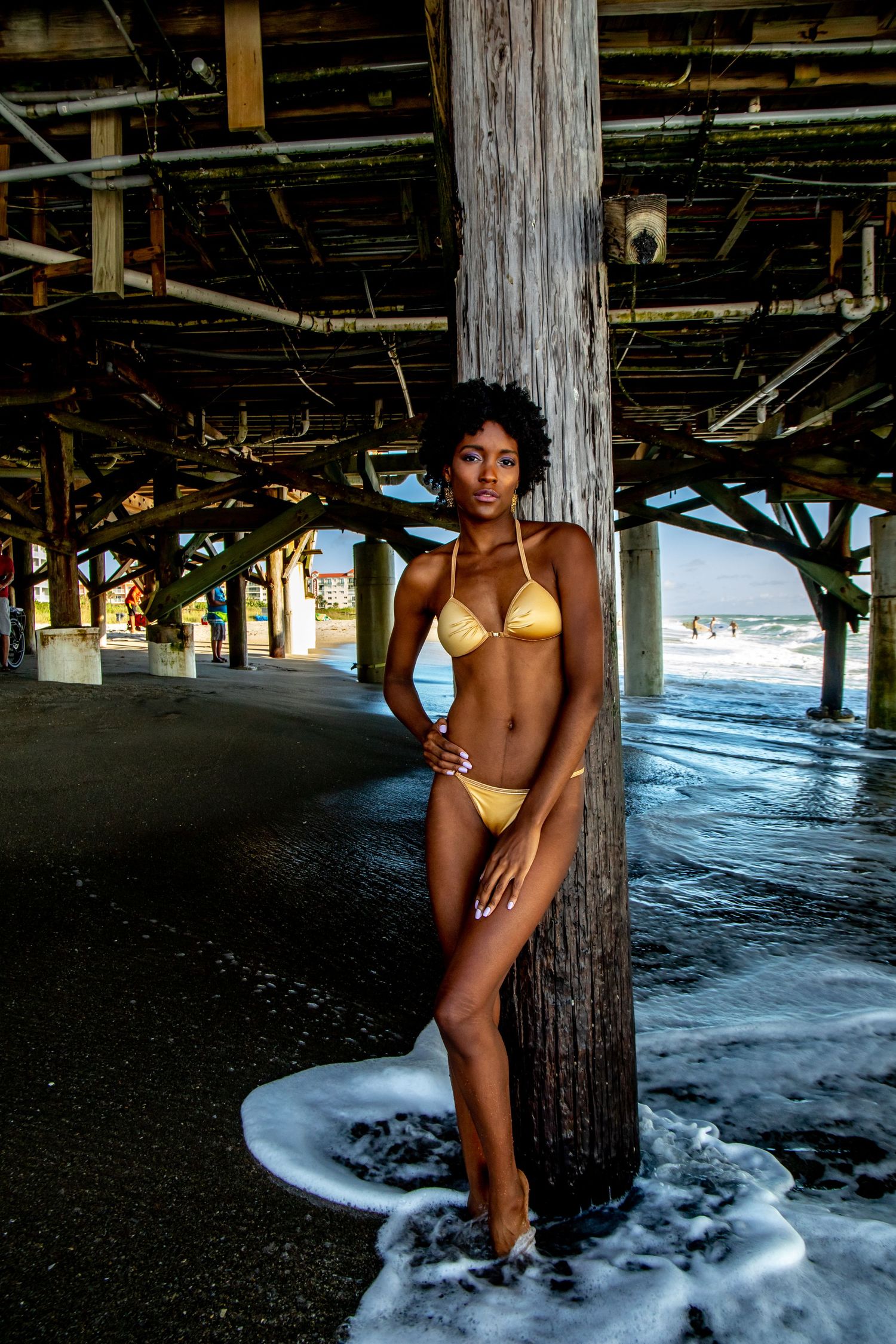 Swimwear model poses beneath weathered wooden pier pilings as ocean waves crash behind.