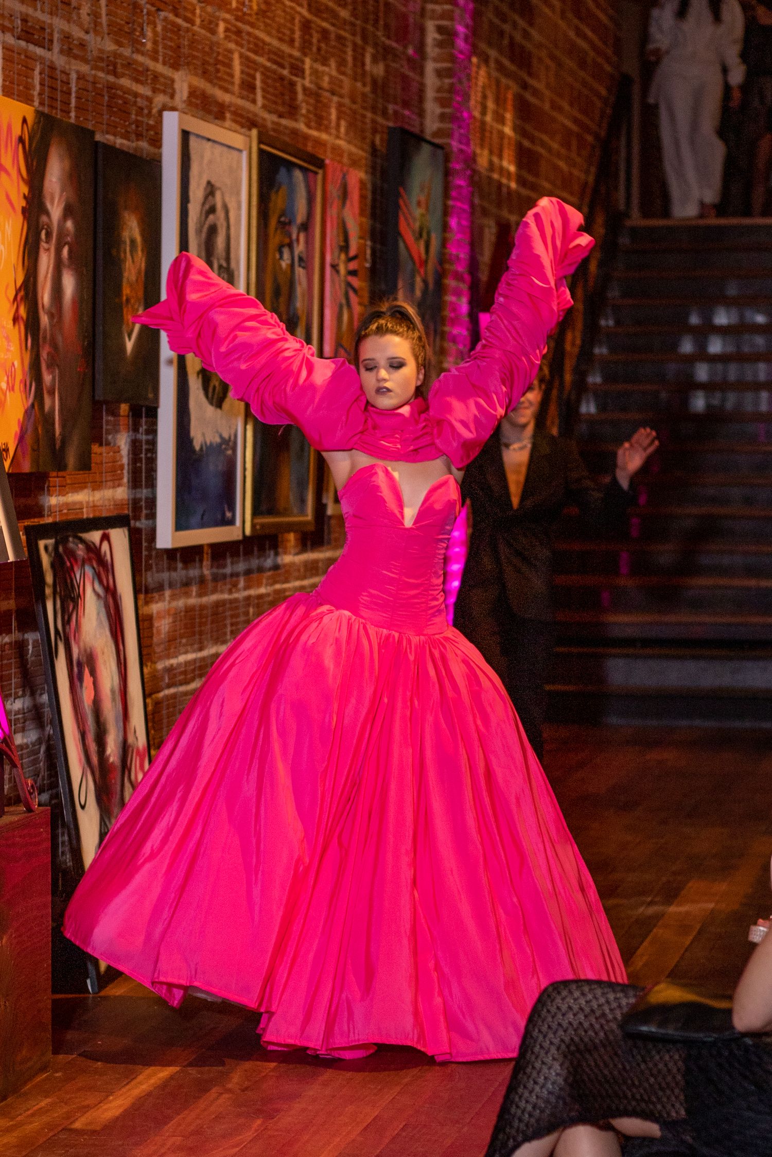 A dramatic fashion show featuring a hot pink ballgown with dramatic sleeves in a brick-walled venue.