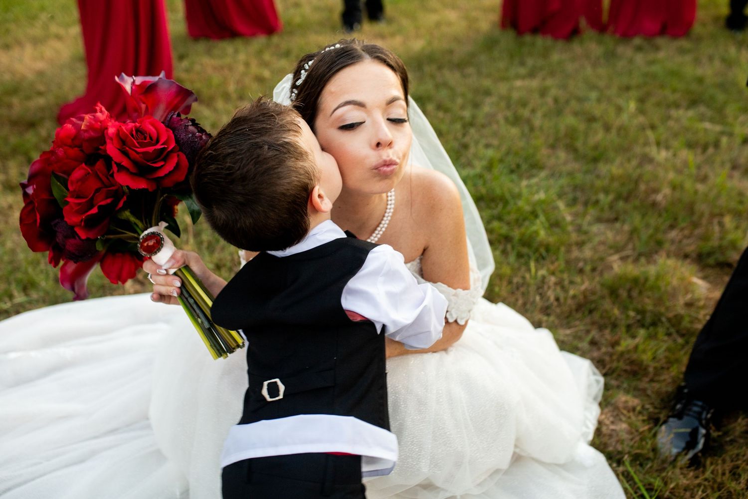 Bride in white gown kneeling to kiss a young ring bearer in black suit holding red roses at outdoor wedding.