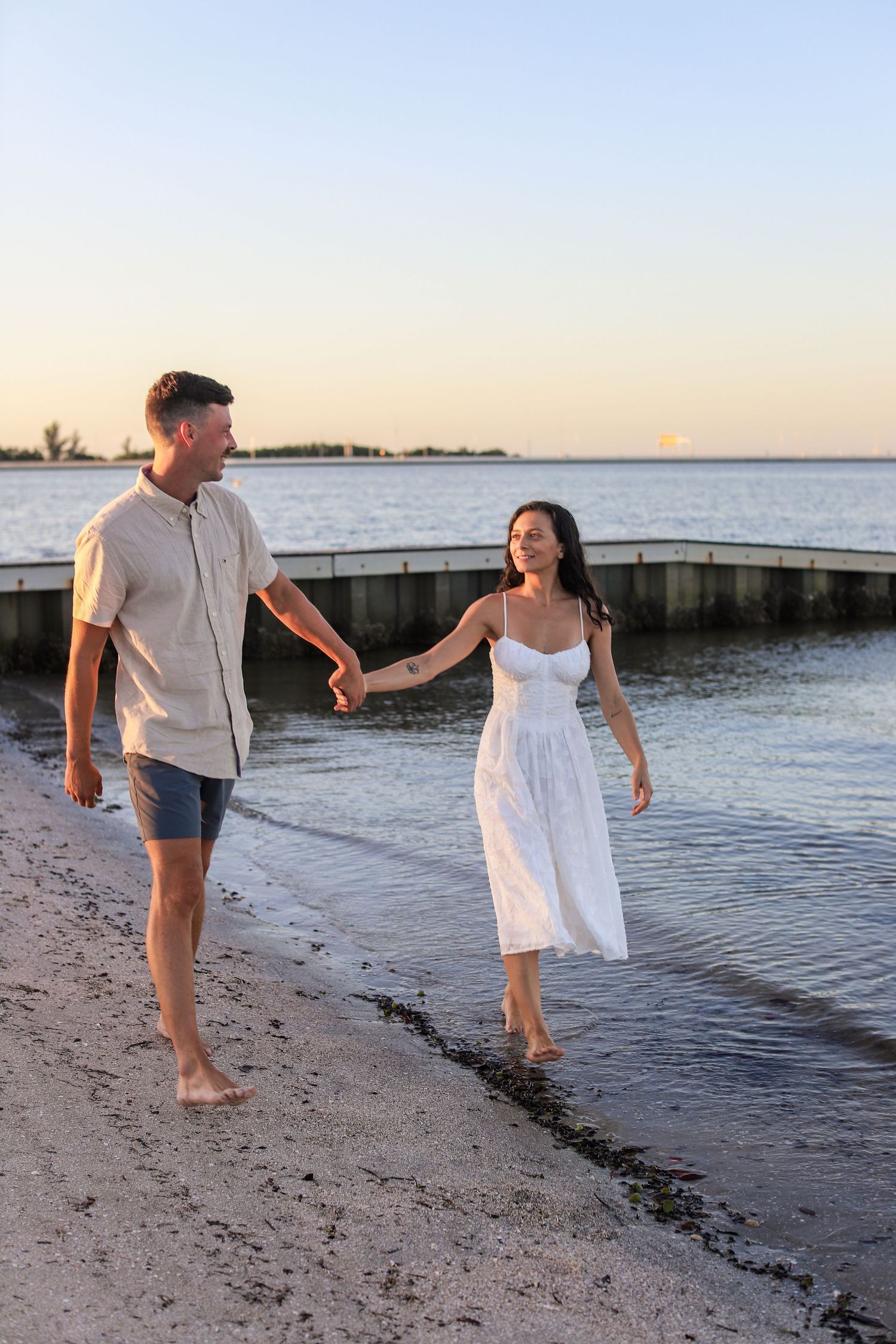 Couple holding hands walking barefoot along a sandy beach at sunset, woman in white dress, man in casual shirt and shorts.