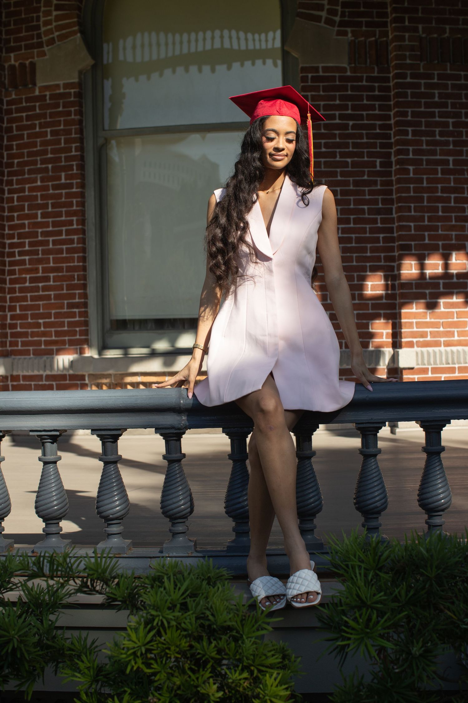 Young woman in white dress and red graduation cap posing on decorative railing outside brick building.
