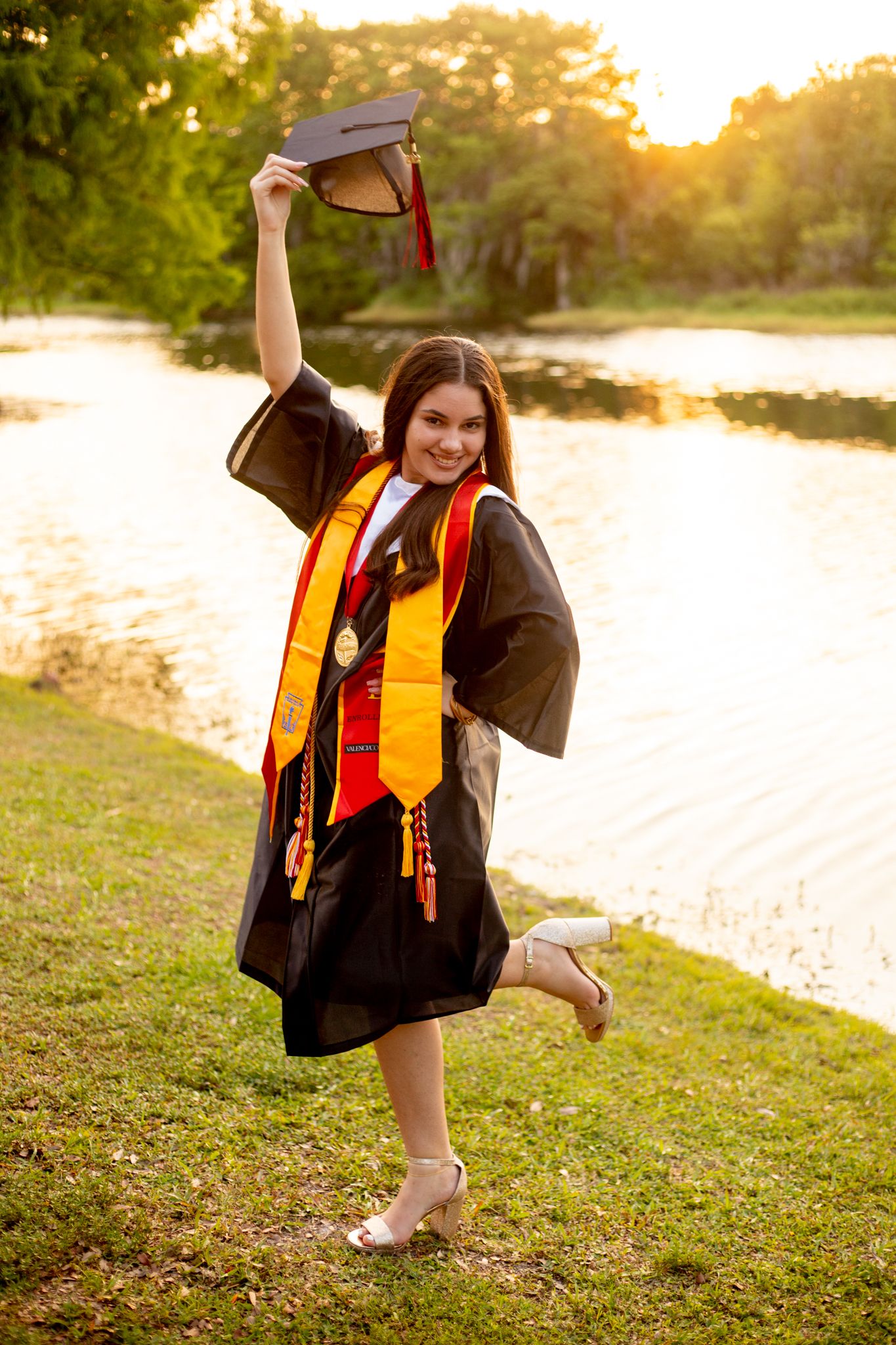 Graduate celebrates by lake at sunset wearing traditional black graduation regalia with gold honors stole.