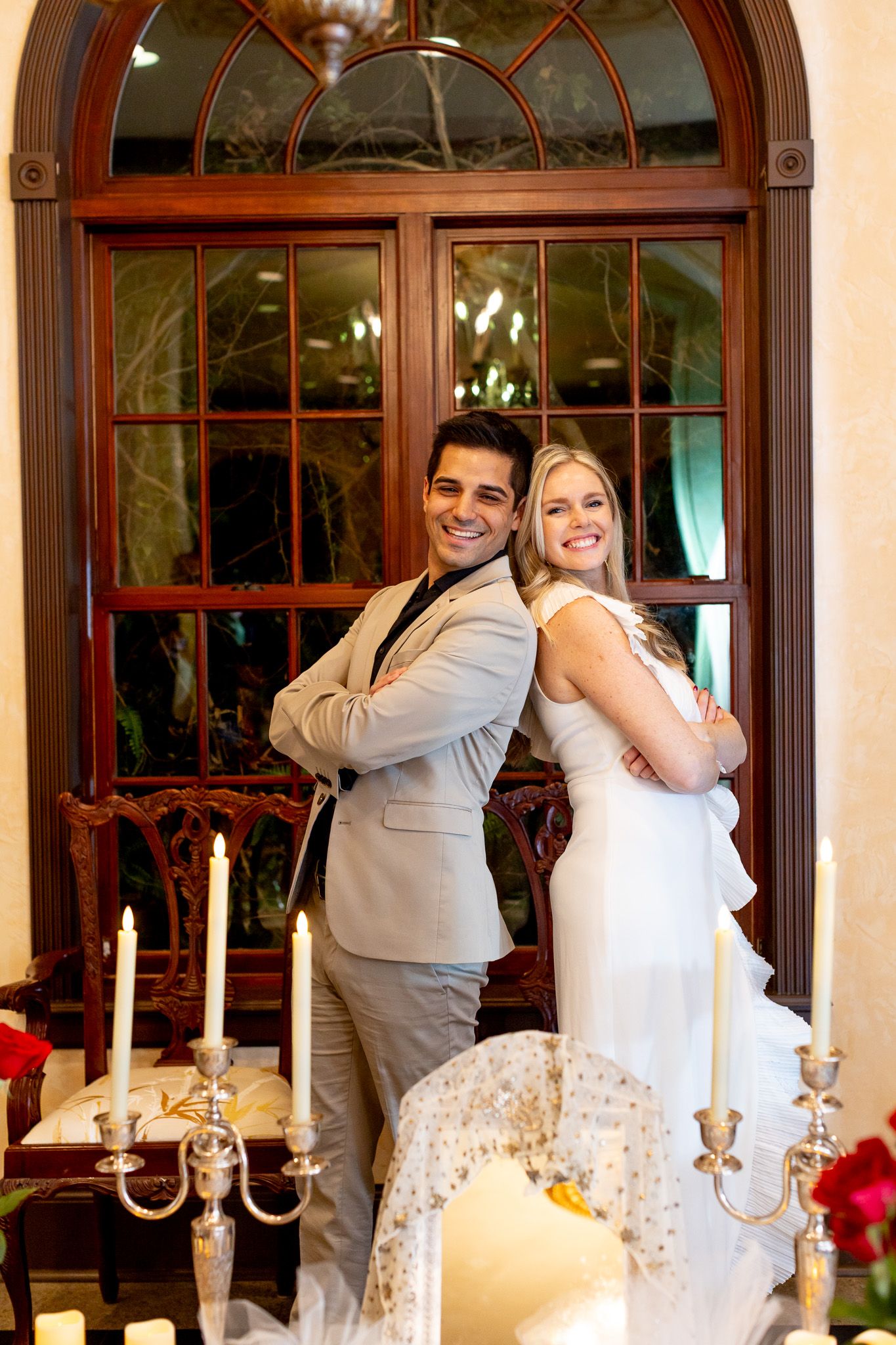 Stylish couple poses back-to-back in formal wear by elegant doorway with candelabras.