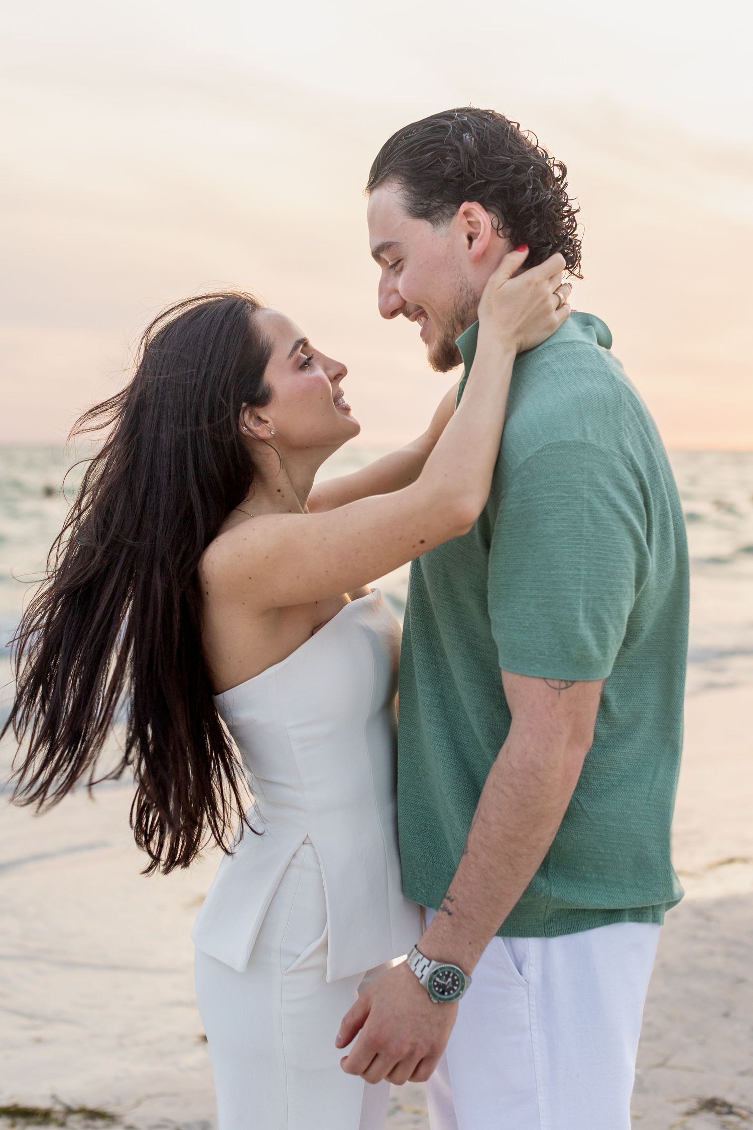A romantic couple embraces on a beach at sunset, woman in white jumpsuit touching man's neck in green shirt.