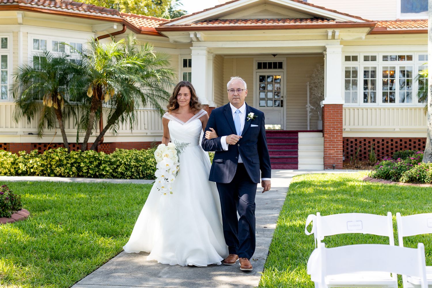 Bride in white gown walks down outdoor aisle with escort in navy suit before a charming Florida wedding venue.