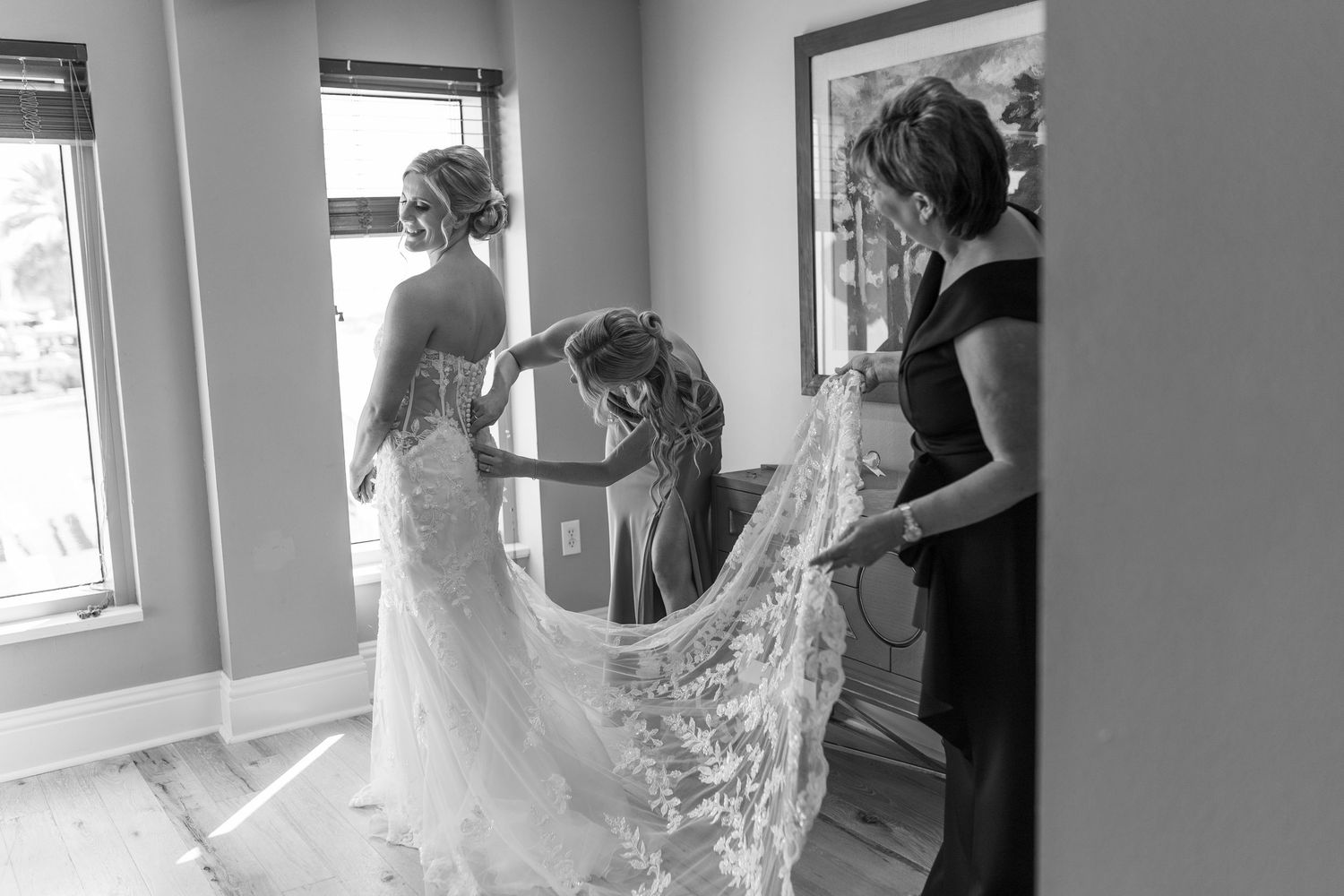 Black and white photo of bride being helped into lace wedding dress by two women in a bright room.