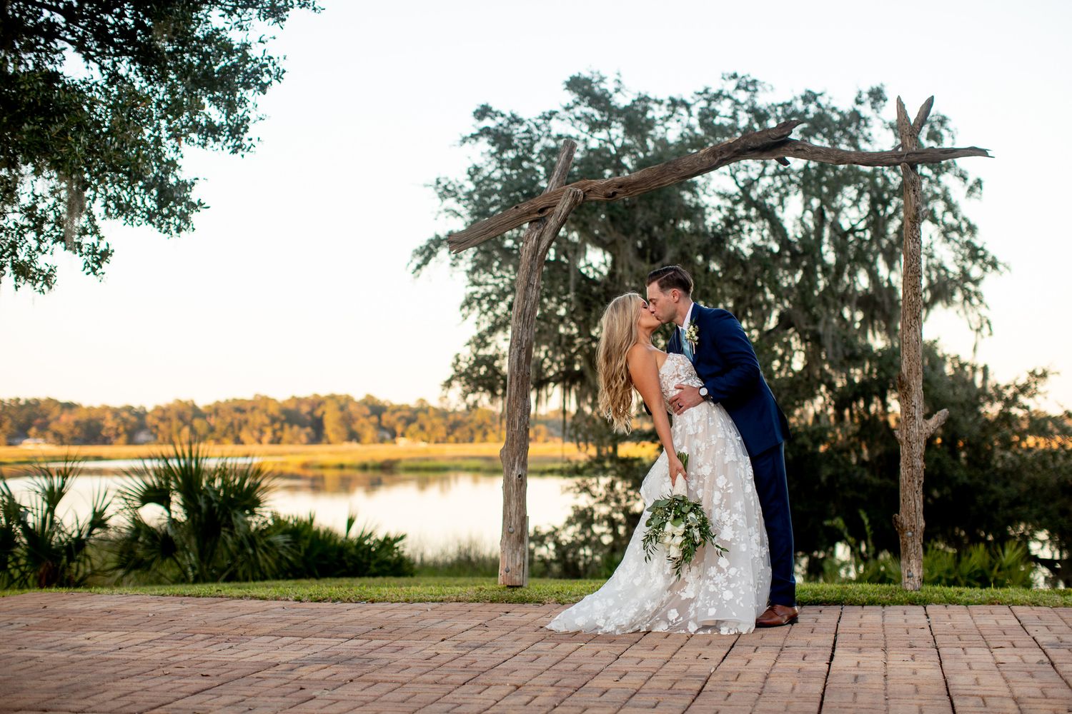 Bride and groom share a romantic dip kiss under a rustic wooden arch by a serene lake at golden hour.