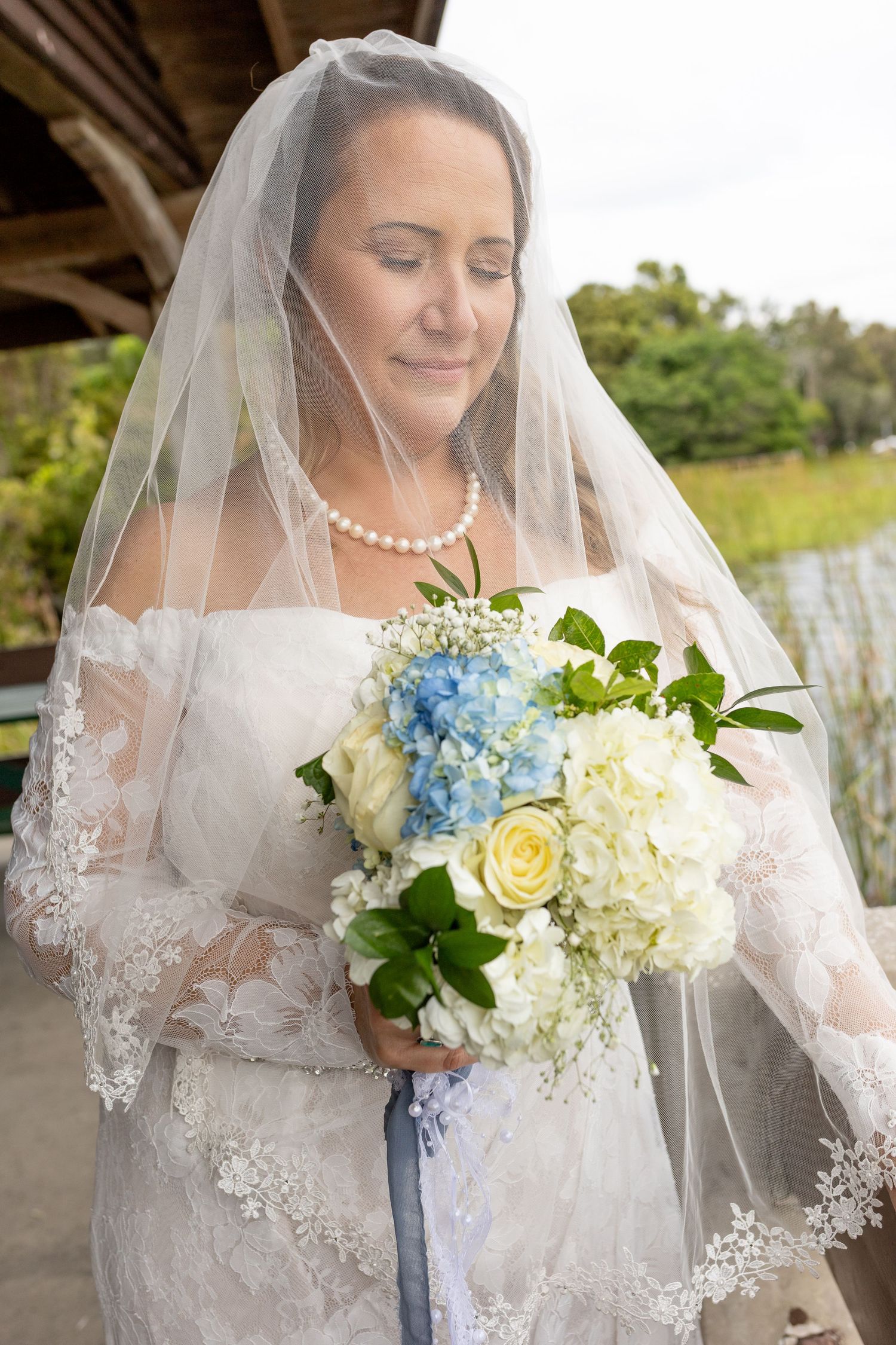 Bride in lace wedding dress and veil holding blue and white floral bouquet with eyes closed outdoors.