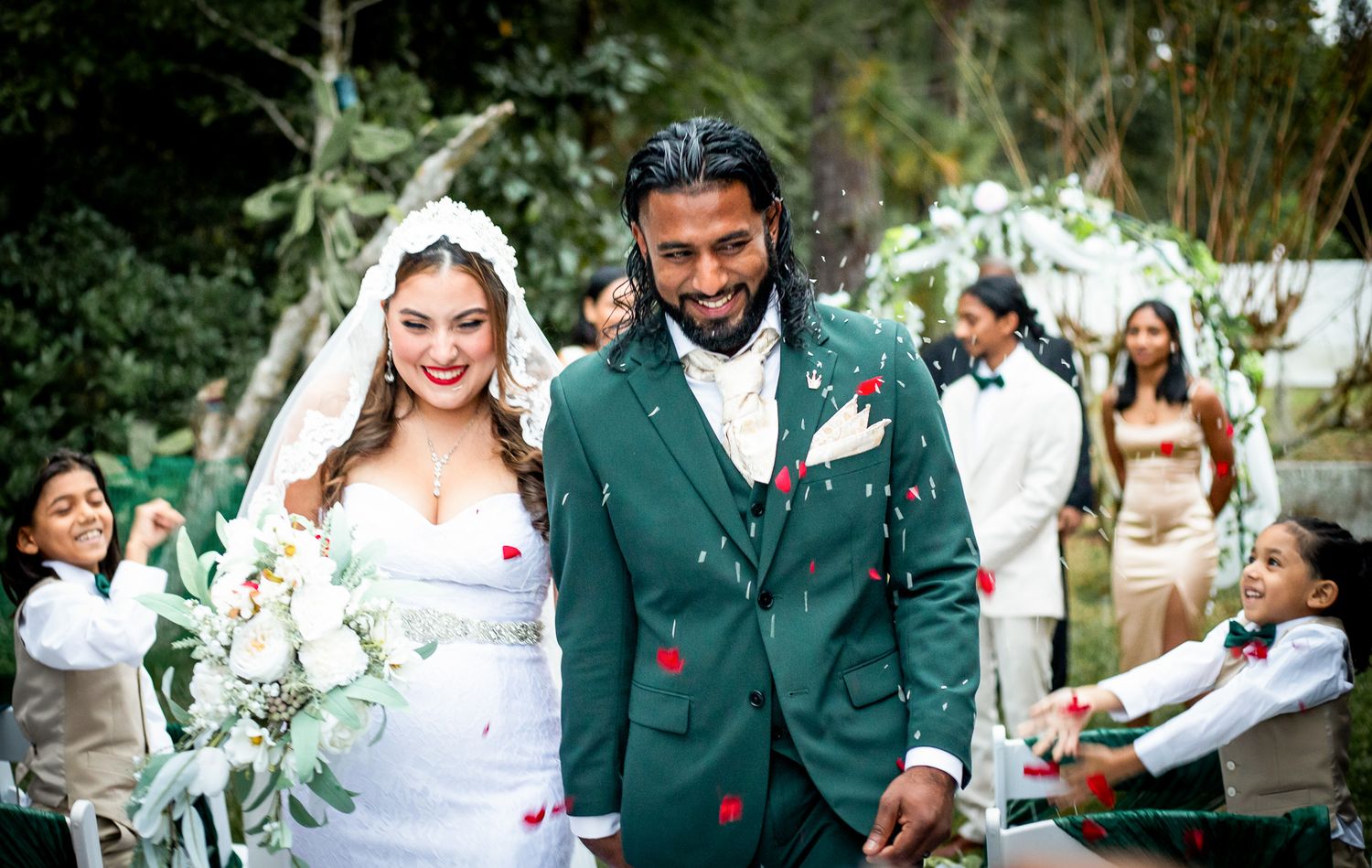 Bride and groom walk down the aisle as guests toss rose petals at outdoor wedding ceremony.
