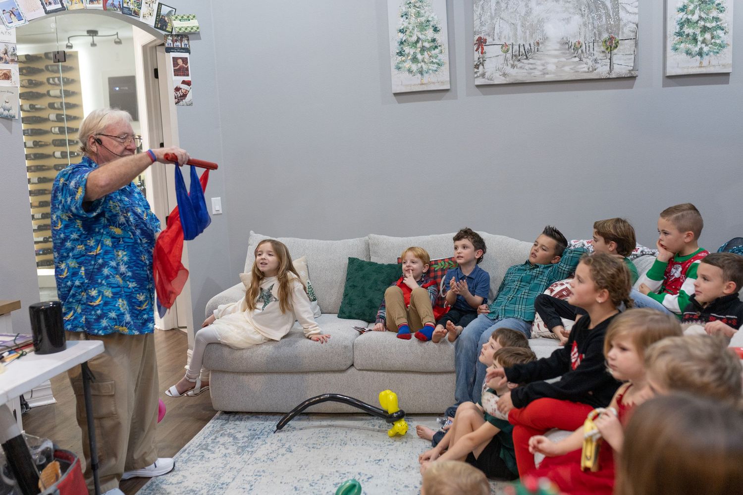 Children gather on couches watching an entertainer in festive attire perform during a holiday celebration.