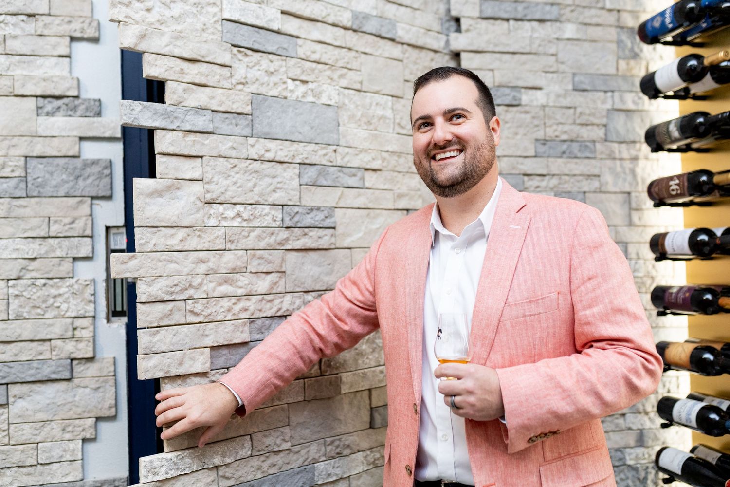 Person in pink blazer smiling while standing next to wine display wall.