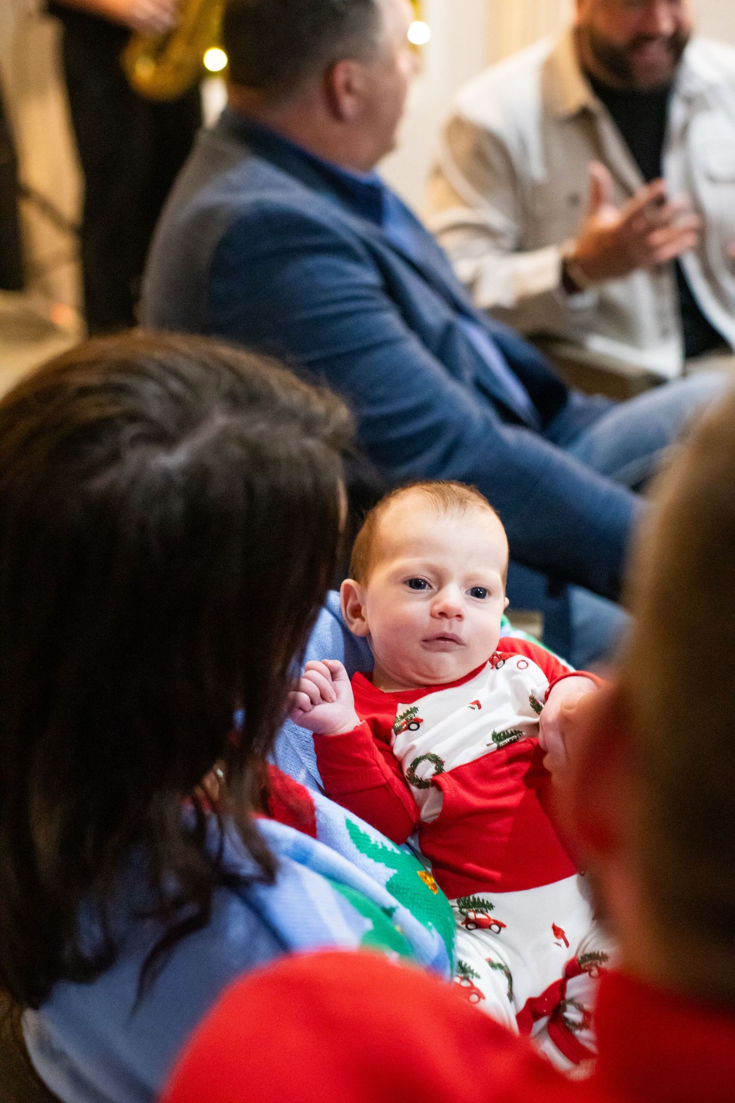 A baby in a red holiday outfit sitting among guests at a Christmas celebration.