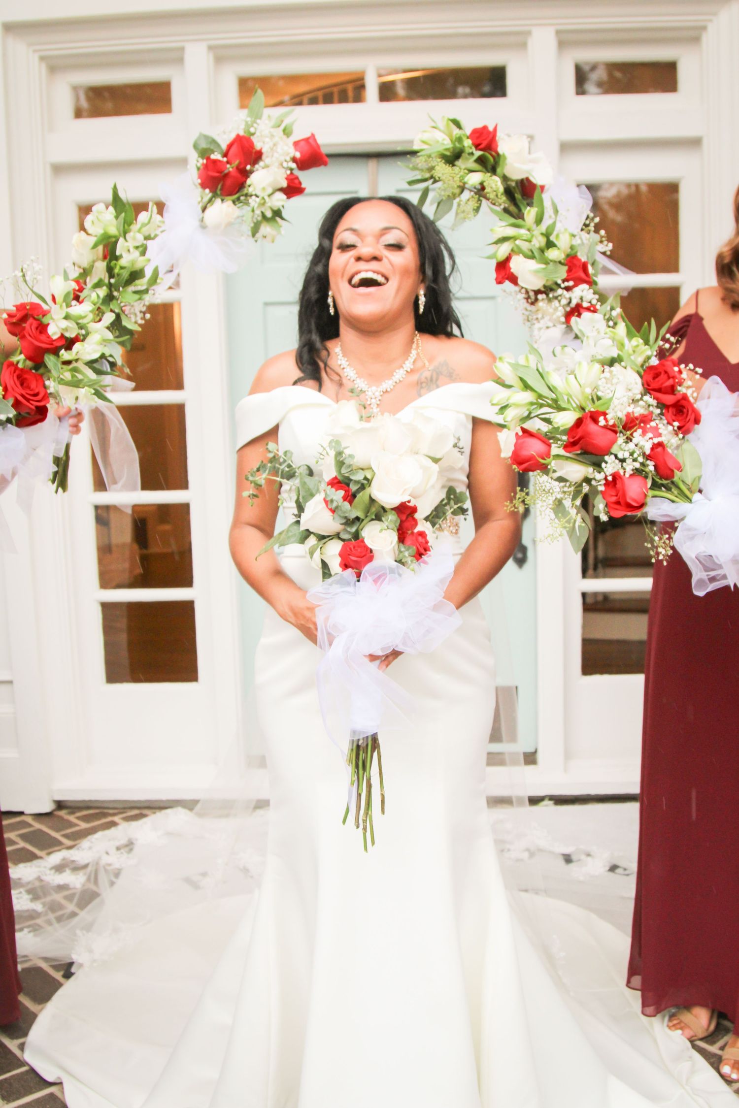 Joyful bride holding bouquet with red and white flowers against white building backdrop.