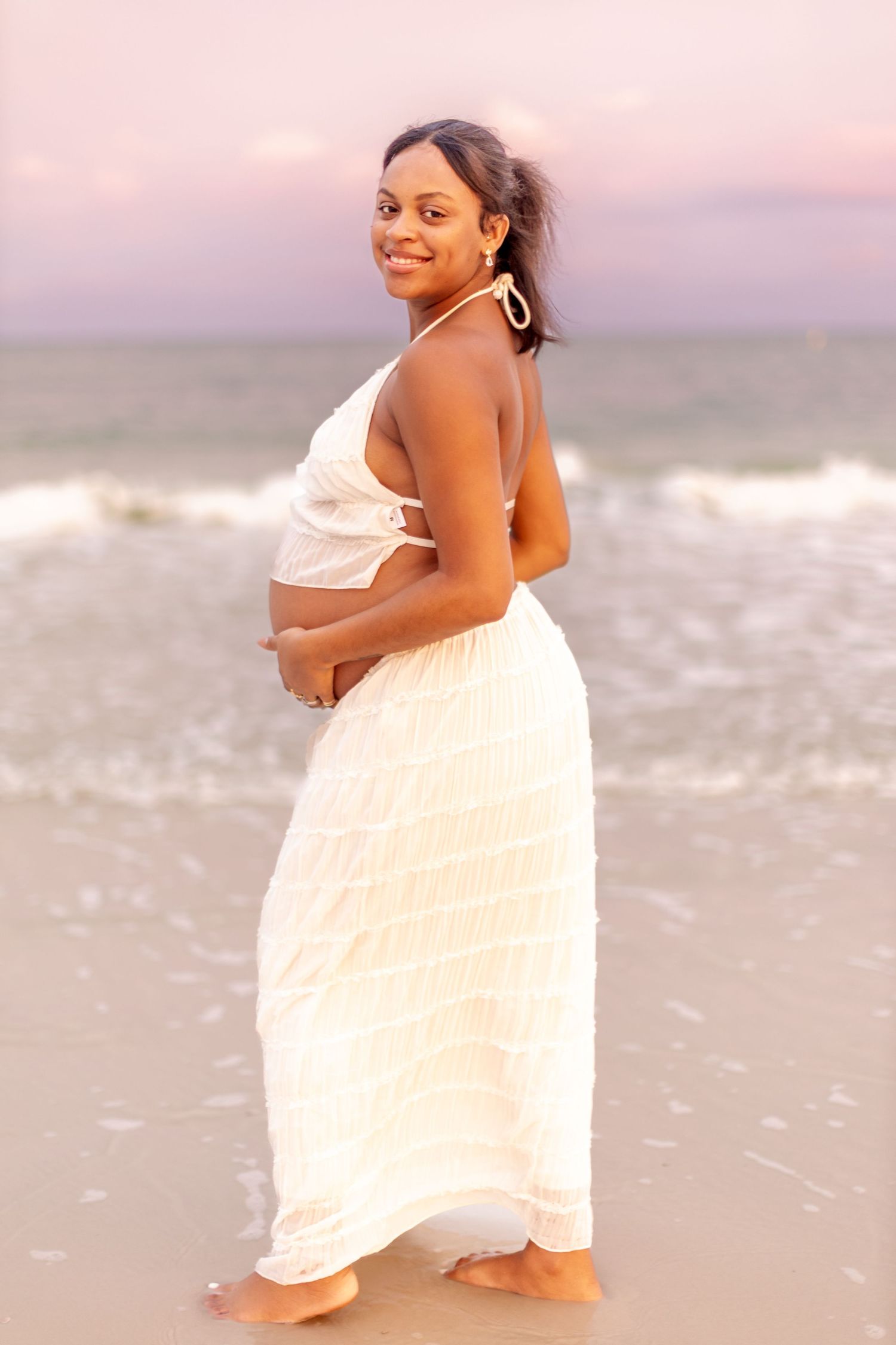 A maternity photoshoot on the beach at sunset shows an expectant mother in a flowing white dress.
