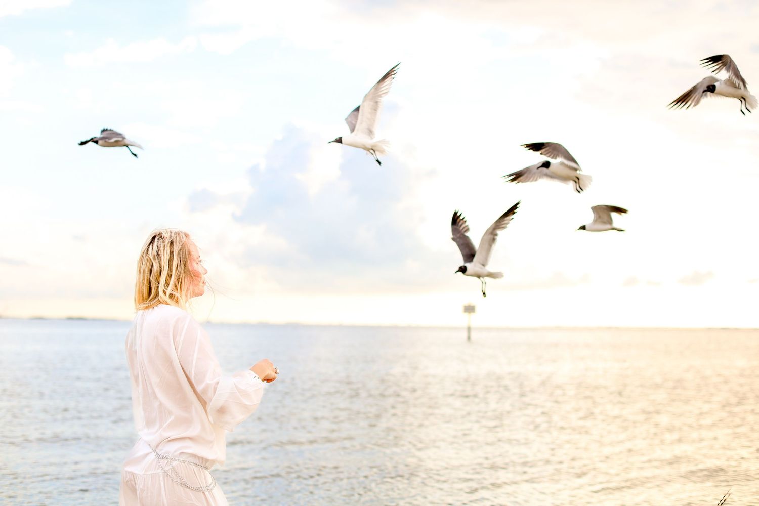 Serene scene of seagulls flying around figure in white by the ocean at sunset.