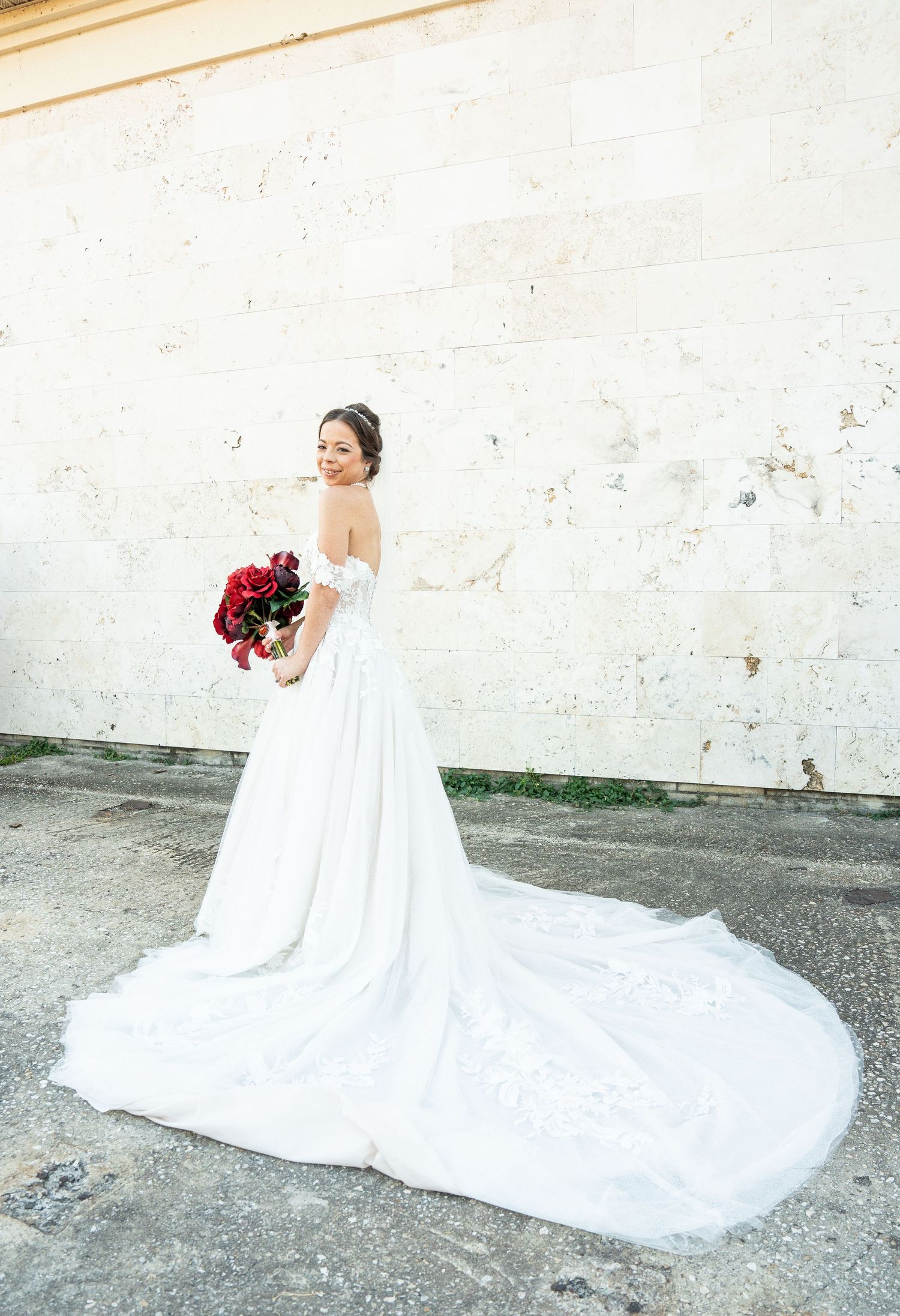Bride in a strapless white ball gown with long train holds red bouquet, posing against a stone wall.