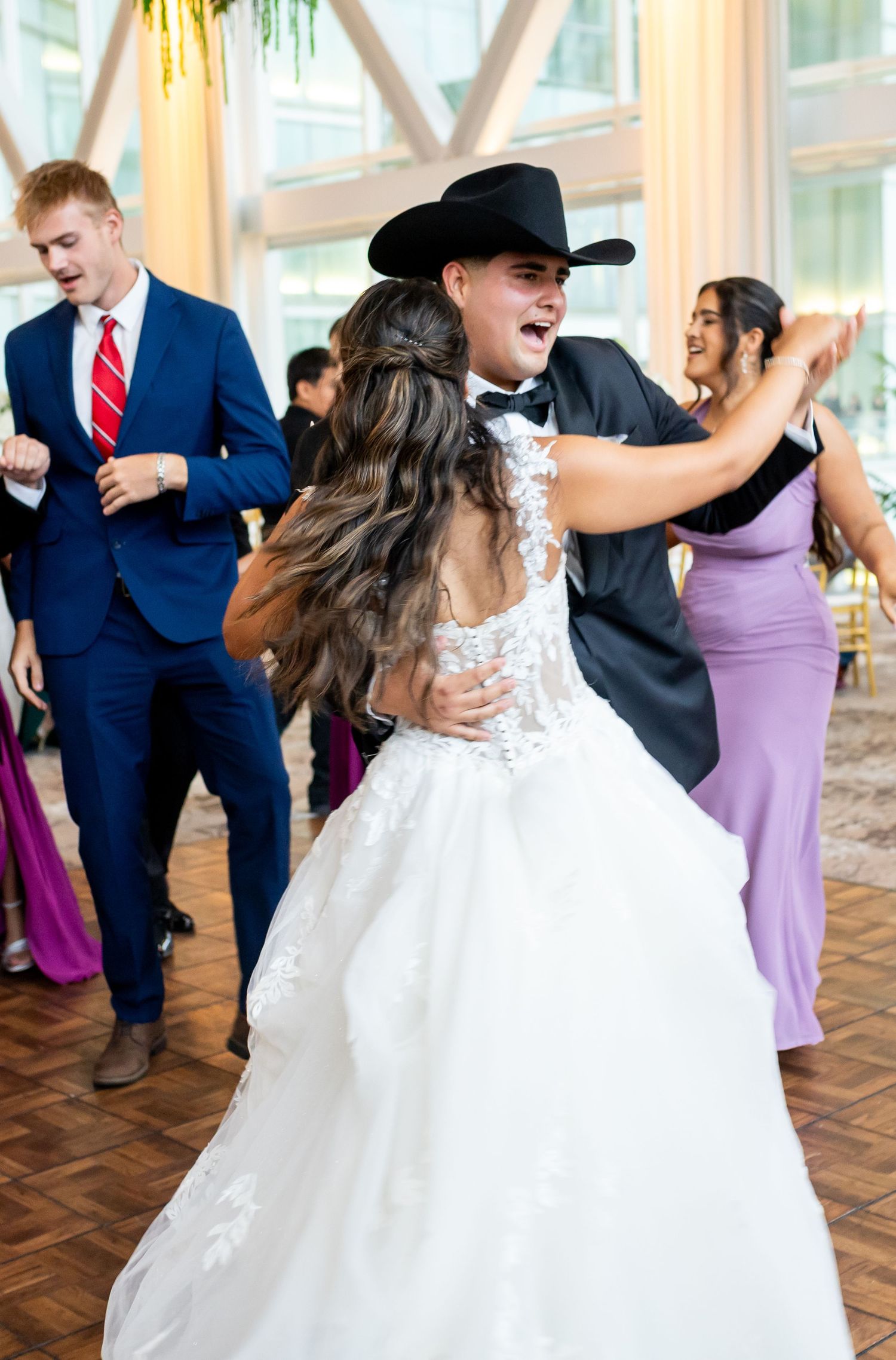 Bride in lace gown dances joyfully with groom in black cowboy hat at elegant wedding reception.