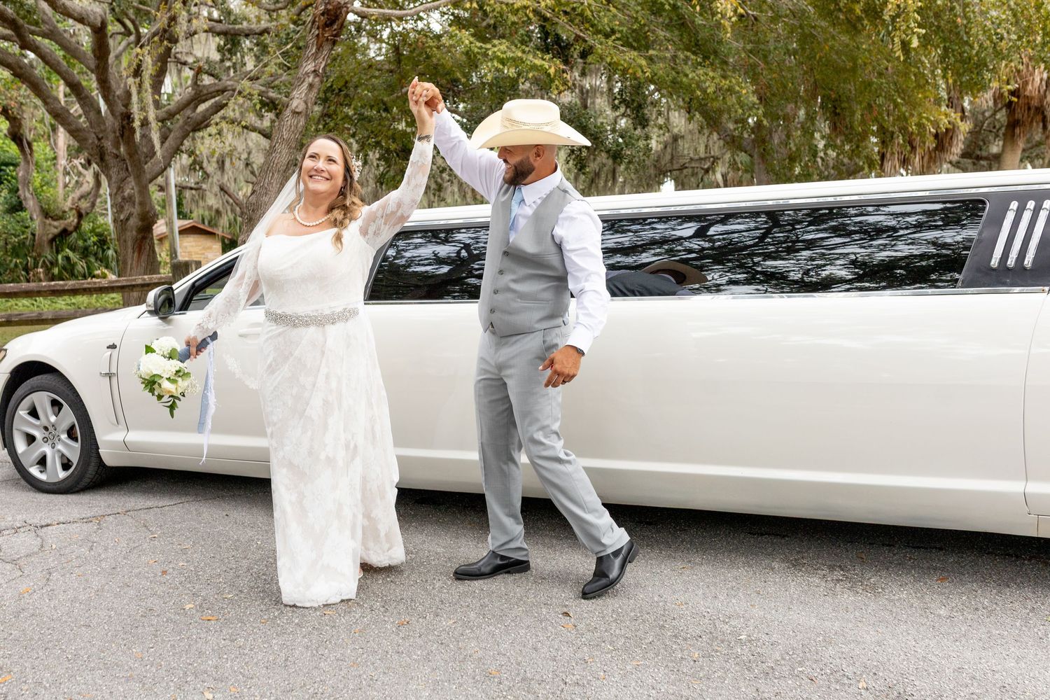 Bride and groom dancing joyfully in front of a white stretch limousine on their wedding day.