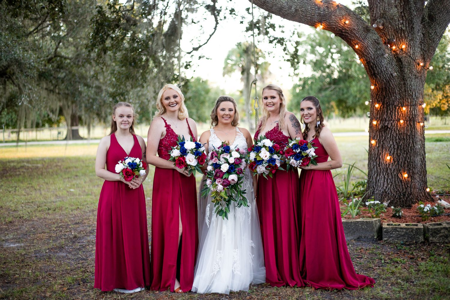 Wedding party in burgundy dresses posing under oak trees at outdoor venue.
