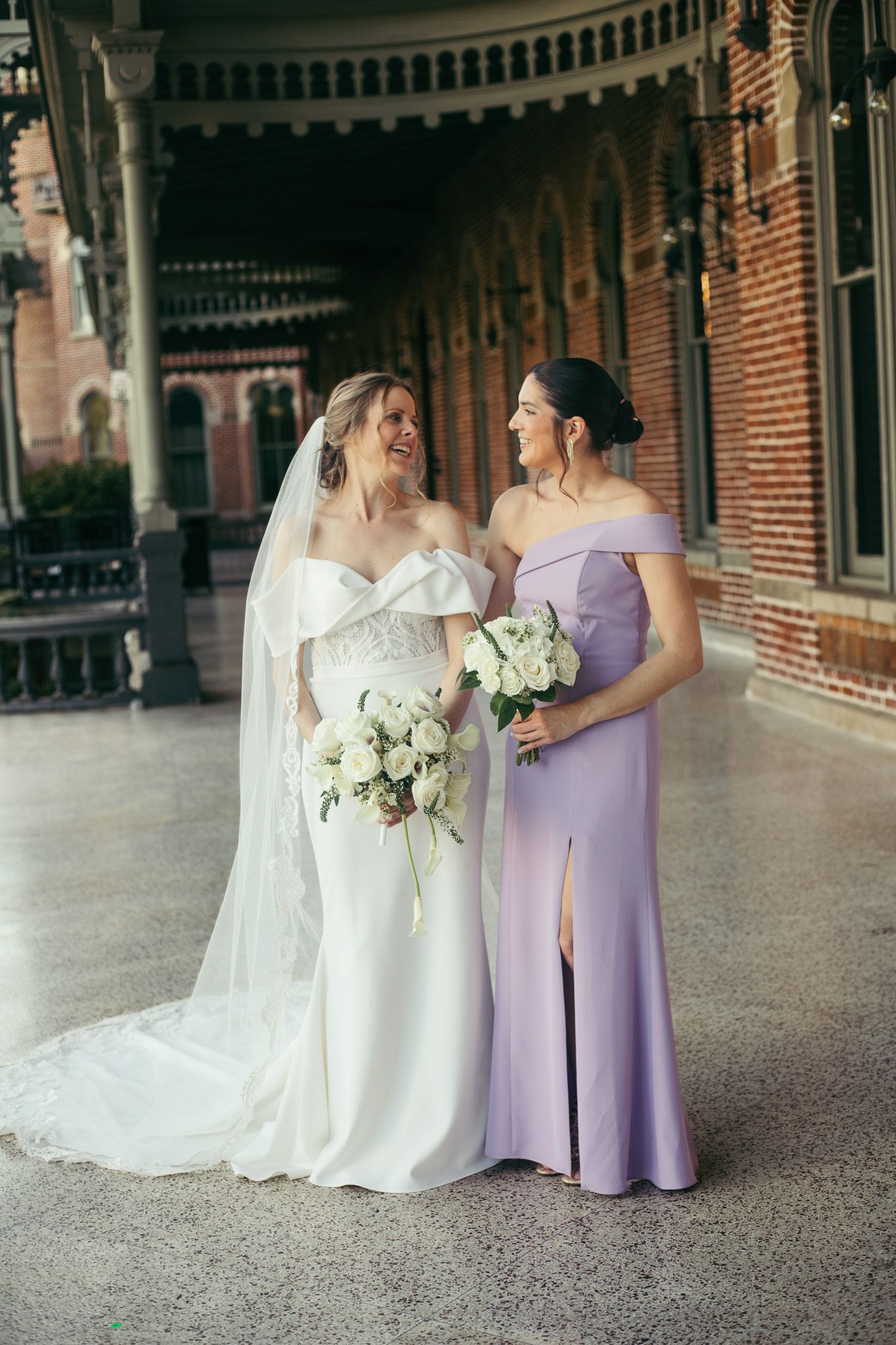 Wedding party in white and lavender dresses posing together in front of historic brick architecture.
