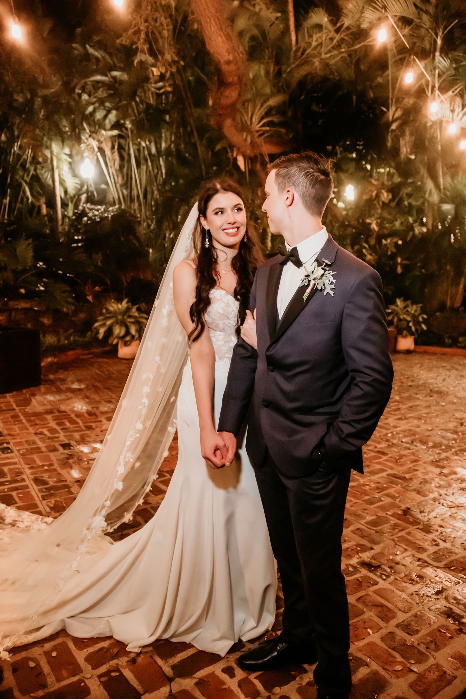 Evening wedding portraits on brick pavers with string lights and tropical foliage creating ambiance.