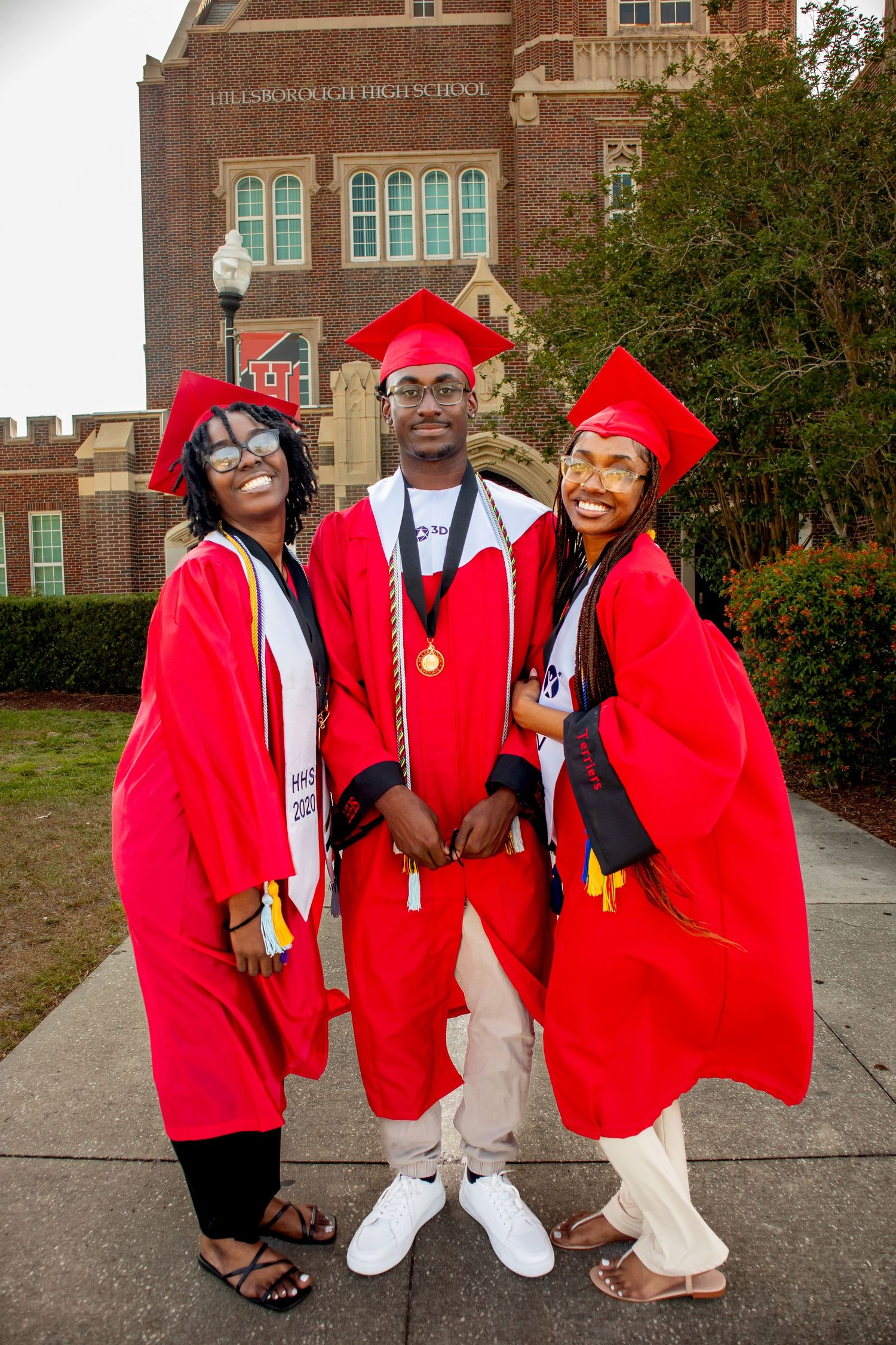 Three graduates pose in bright red graduation regalia outside traditional brick school building.