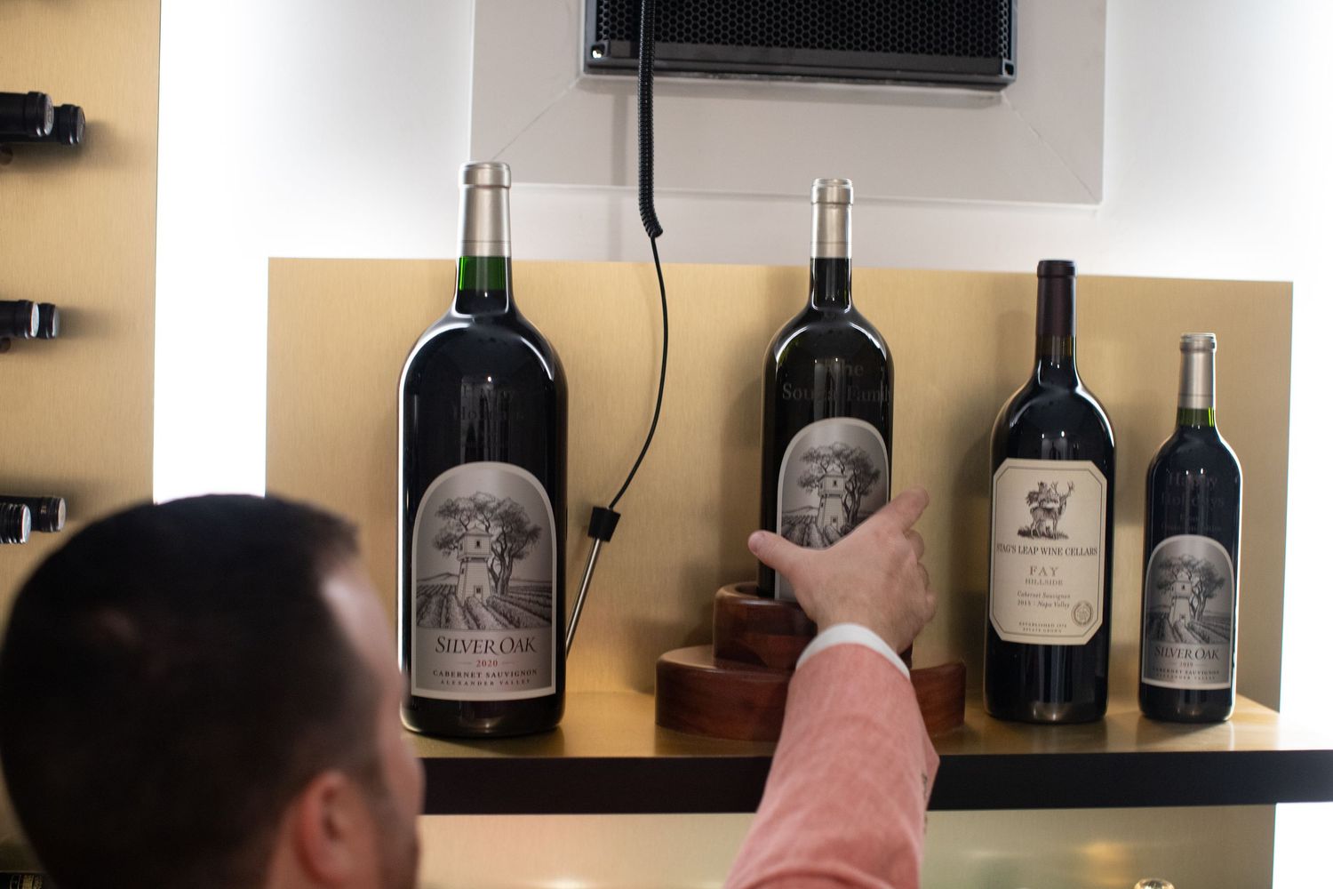 A hand points to wine bottles displayed on a wooden shelf mounted on a wall.