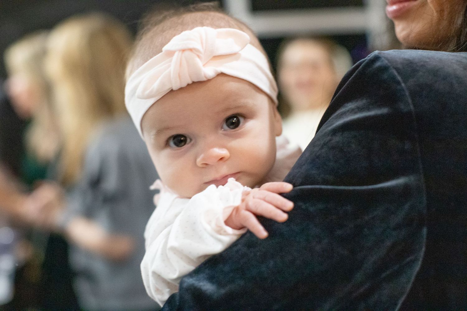 Baby in white outfit being held during a celebration.