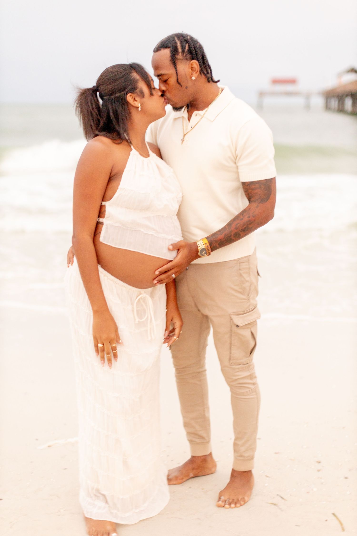 A couple shares a tender moment on the beach wearing matching white and beige outfits.