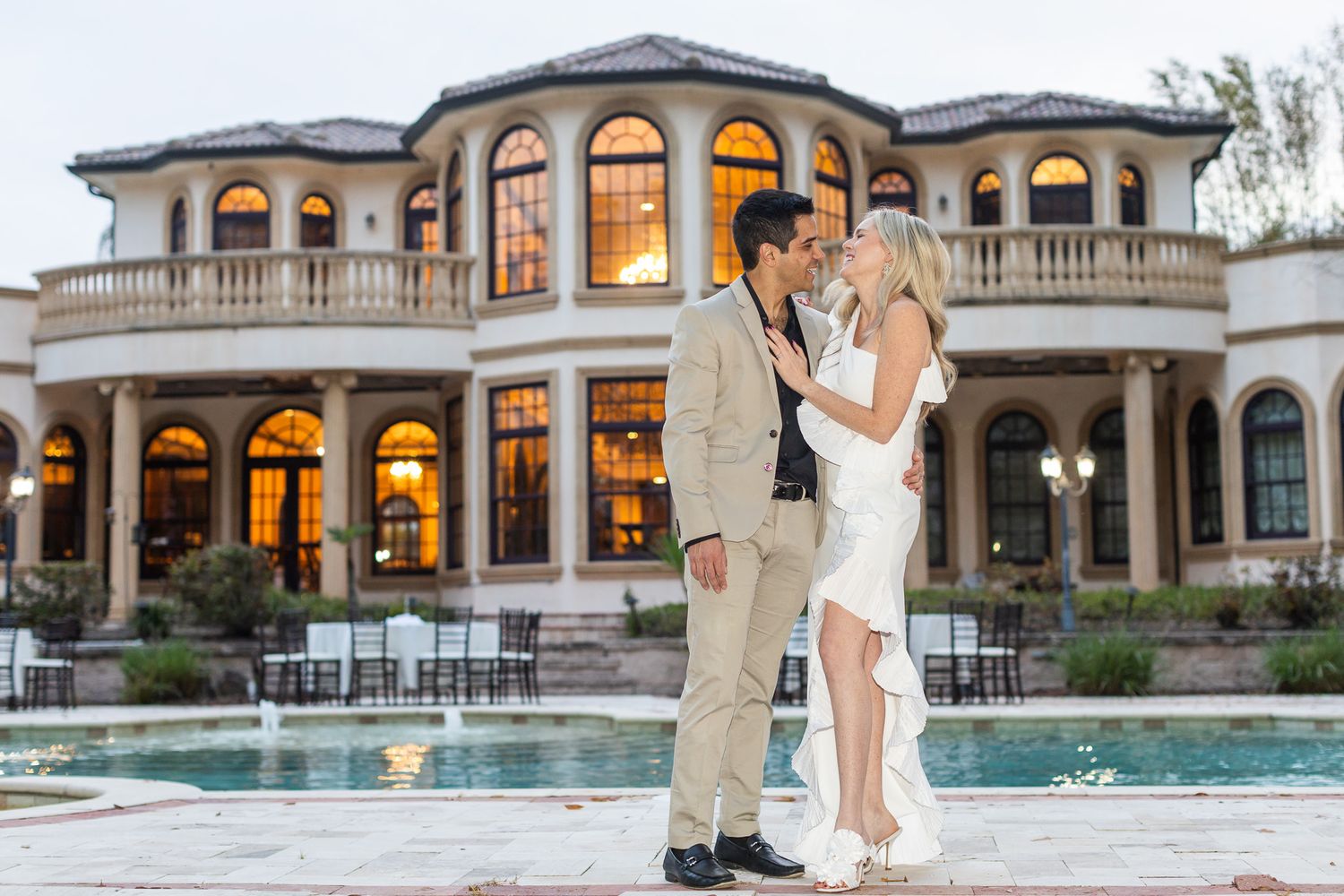 Elegant couple poses in front of luxury mansion with lit pool at dusk.