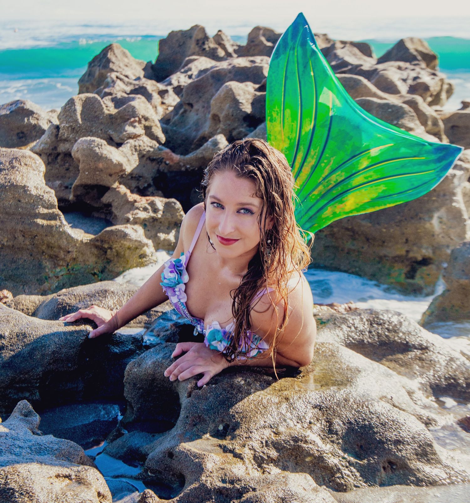 Beachside photoshoot featuring a mermaid tail costume against rocky coastal scenery.