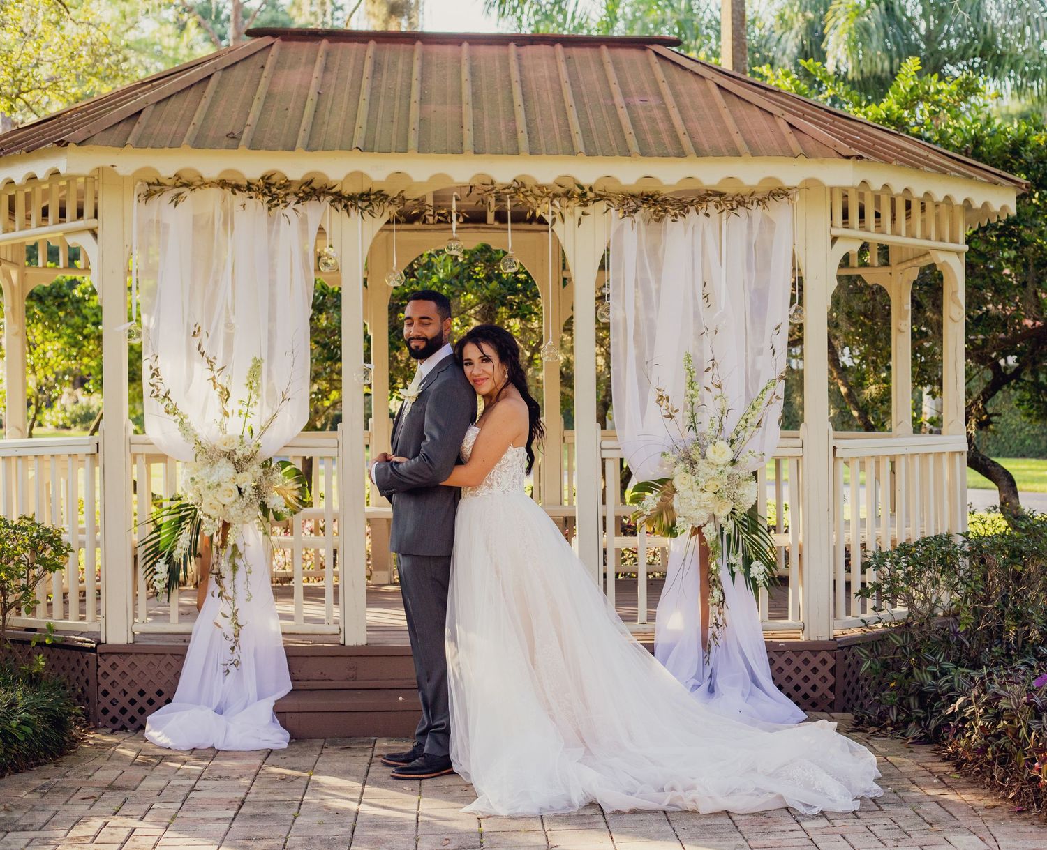 A romantic wedding ceremony under a decorated gazebo with white draping and floral arrangements.