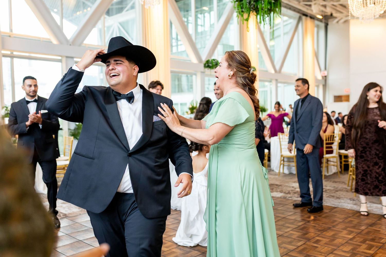 A groom in a black tuxedo and cowboy hat dances joyfully with a woman in a sage green gown at a wedding reception.