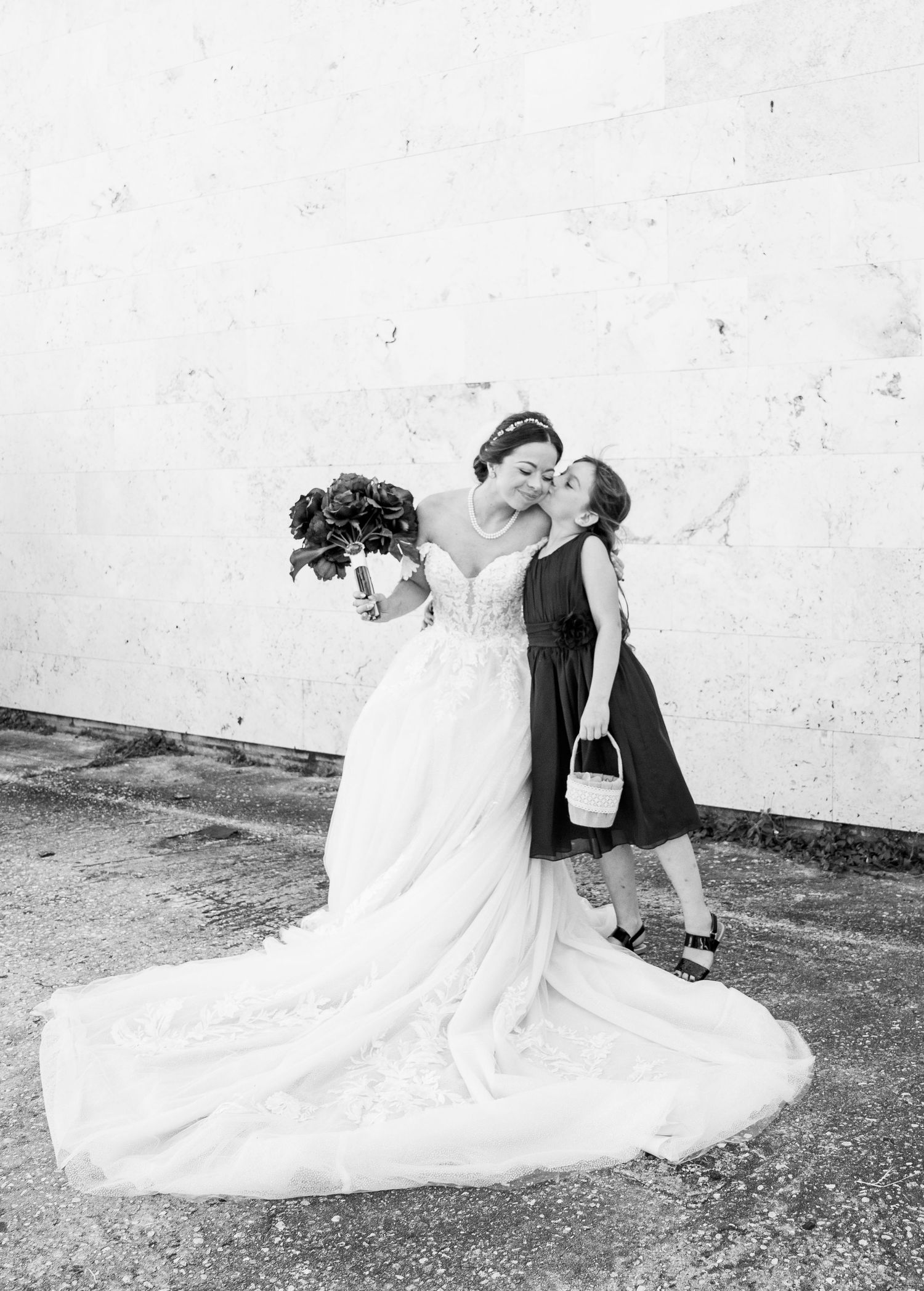 Black and white photo of a bride in a white gown holding bouquet while a young flower girl kisses her cheek.