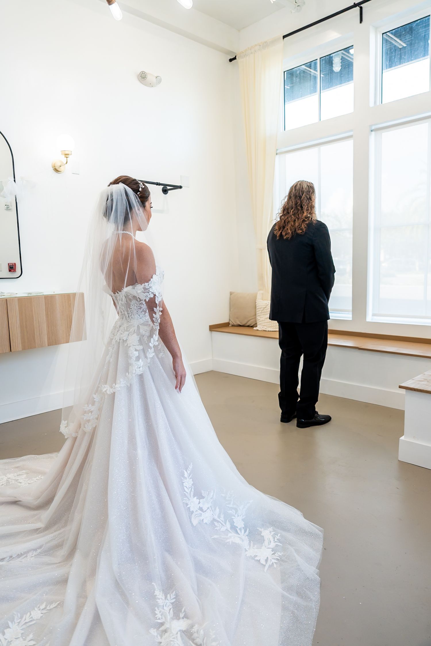 Bride in a lace ball gown with long train and veil stands behind groom facing window for first look.