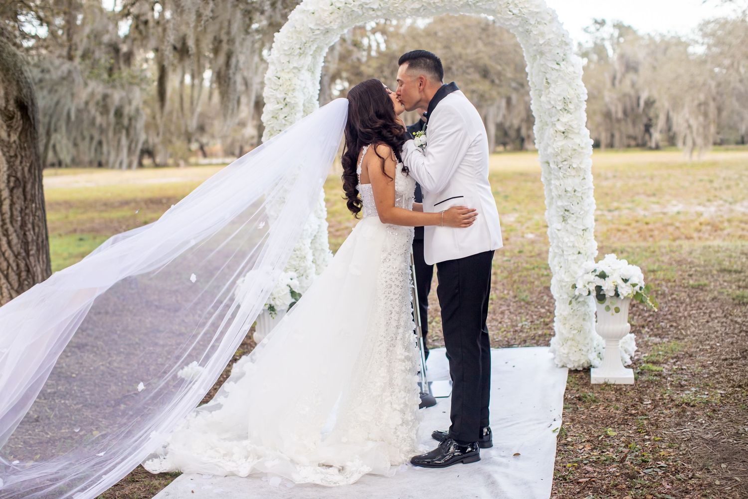 An outdoor wedding ceremony setup with white chairs and decorative columns under Spanish moss trees.