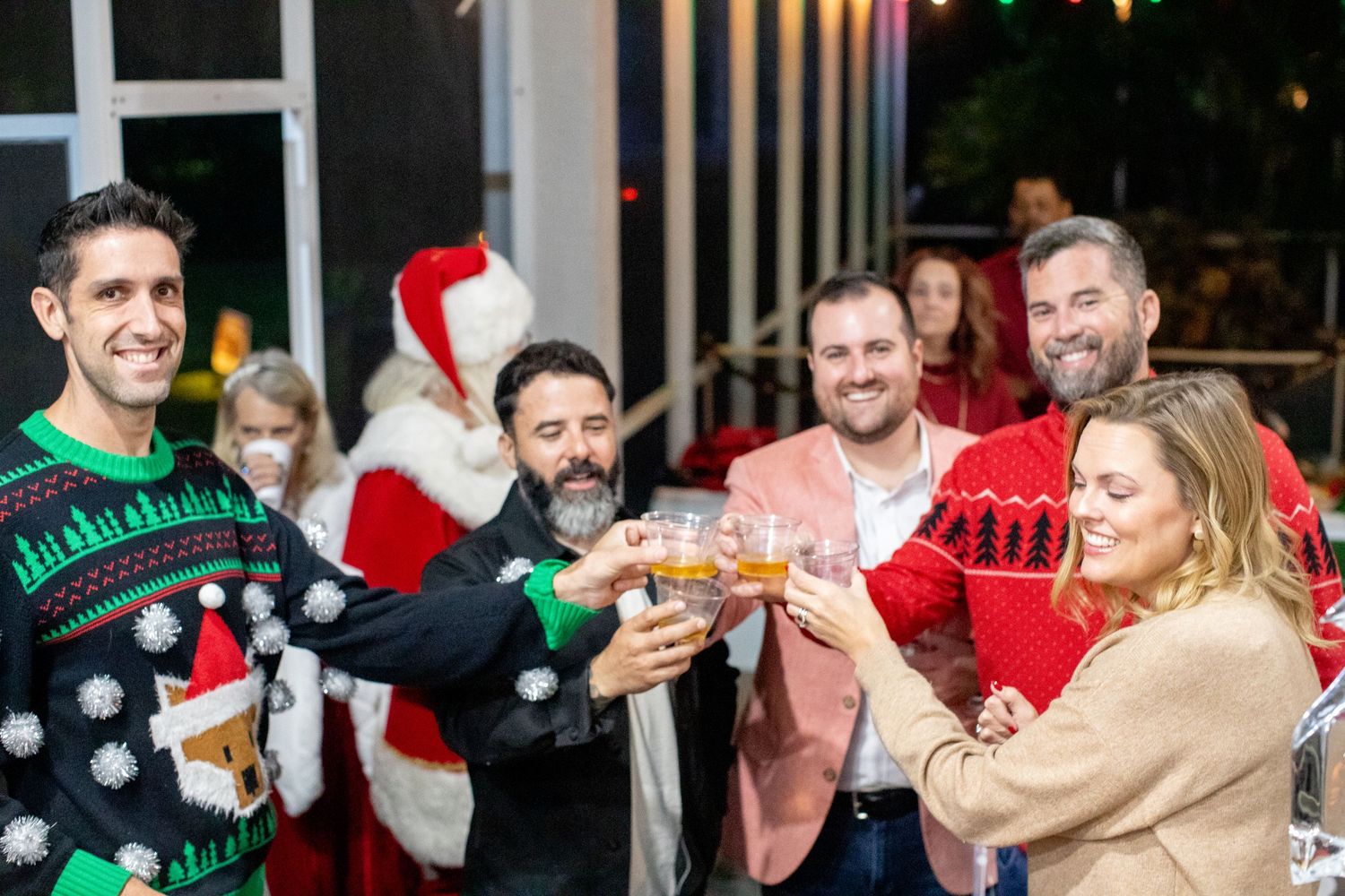 Friends in Christmas sweaters toast and celebrate at a holiday party with Santa in the background.